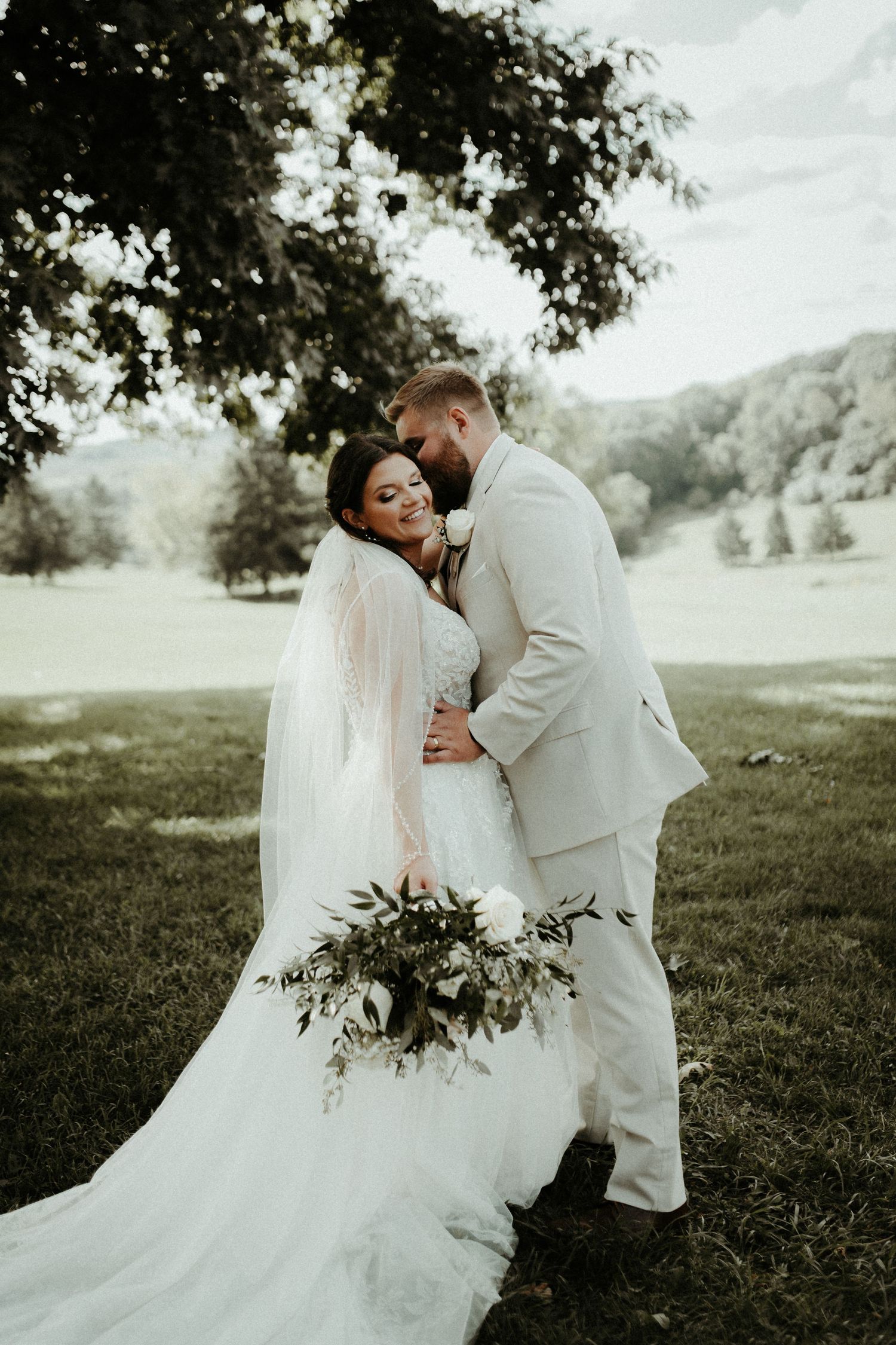 Newlywed couple embraces outdoors in a romantic wedding photo with a beautiful bouquet against a pastoral backdrop. Pittsburgh Wedding