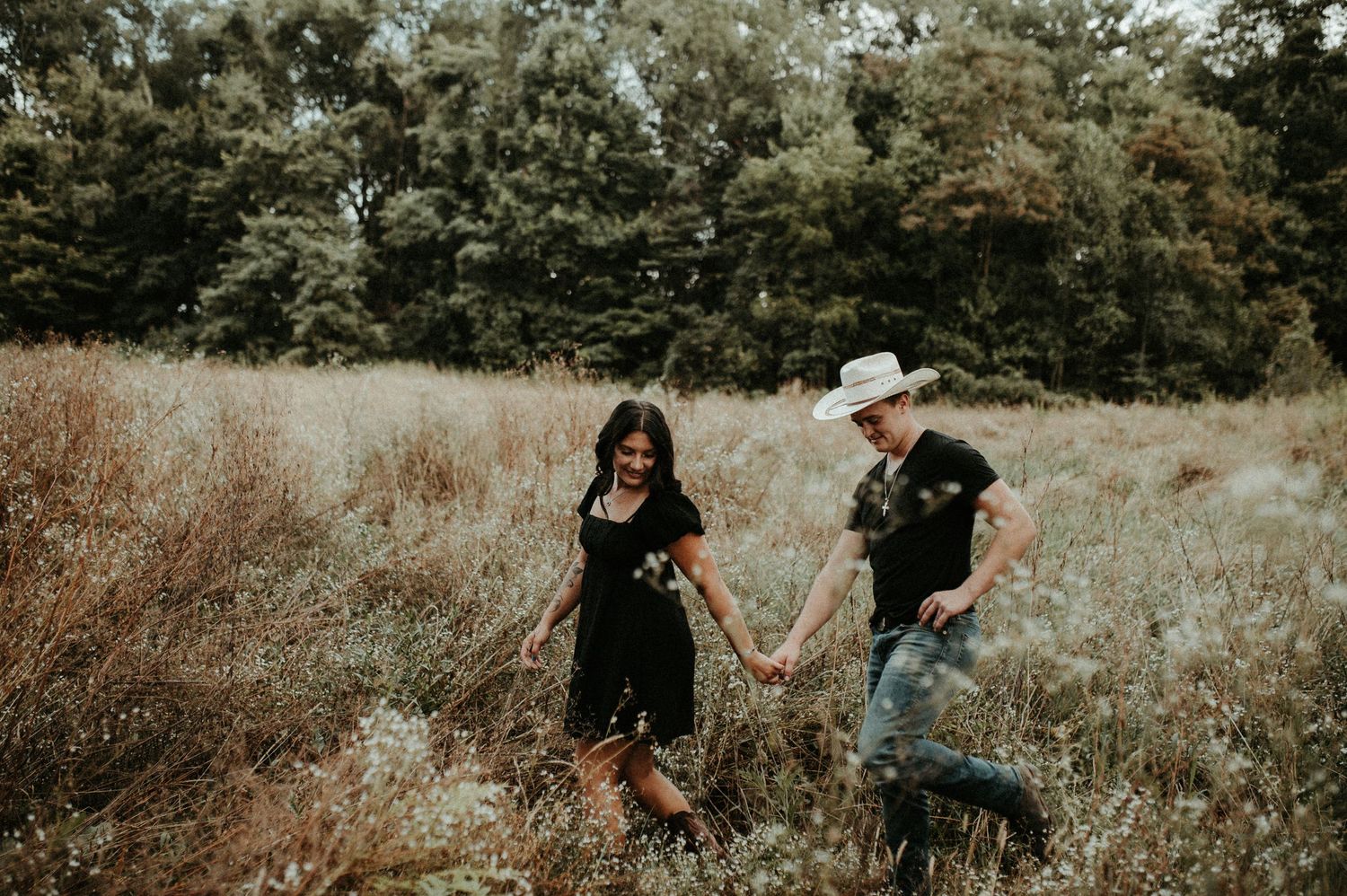 Couple holding hands while walking through a rustic field with tall grass and trees in the background. Pittsburgh Wedding Photographer