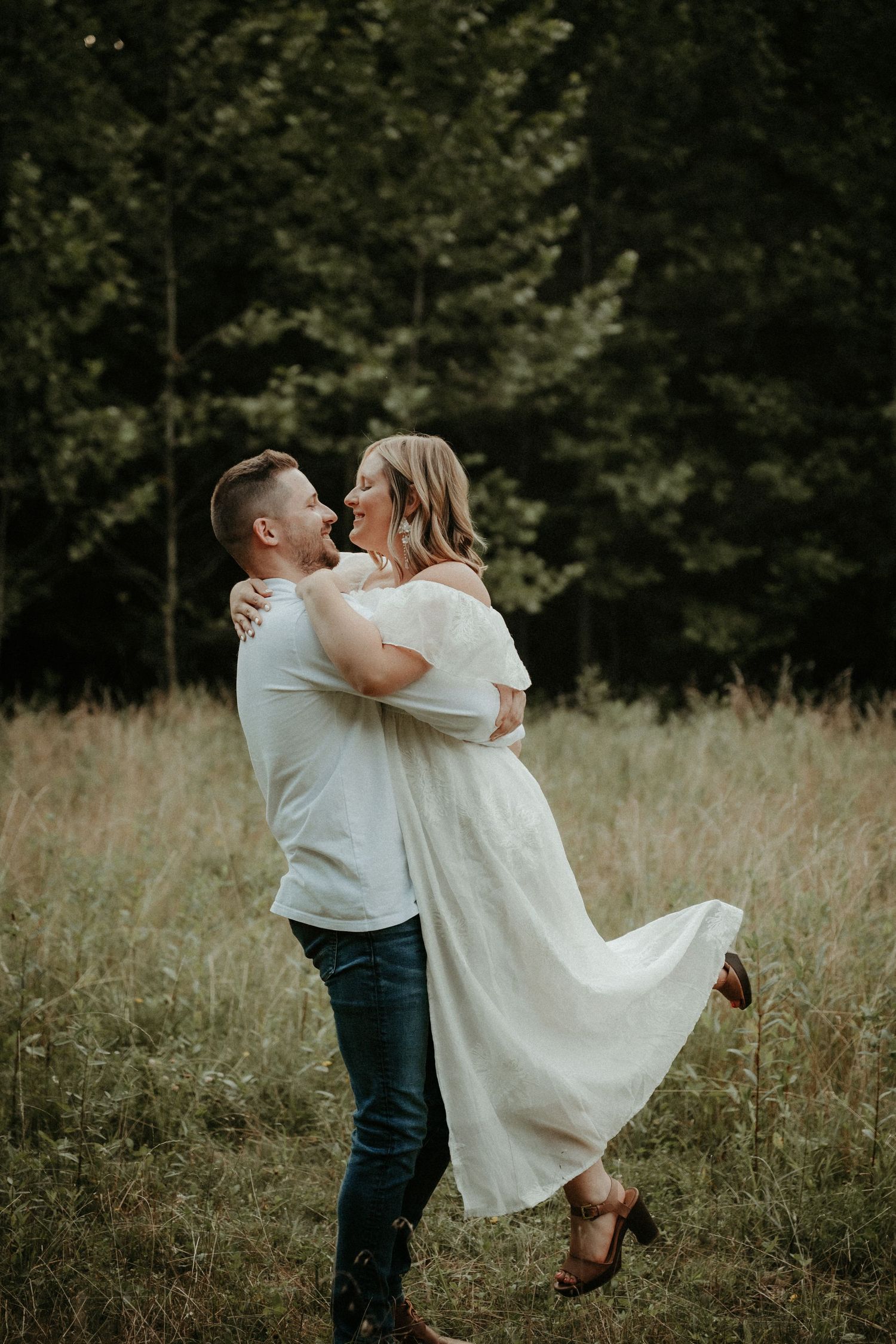 A romantic couple embraces in a sunlit meadow, with the person in white being lifted and twirled against a backdrop of trees.