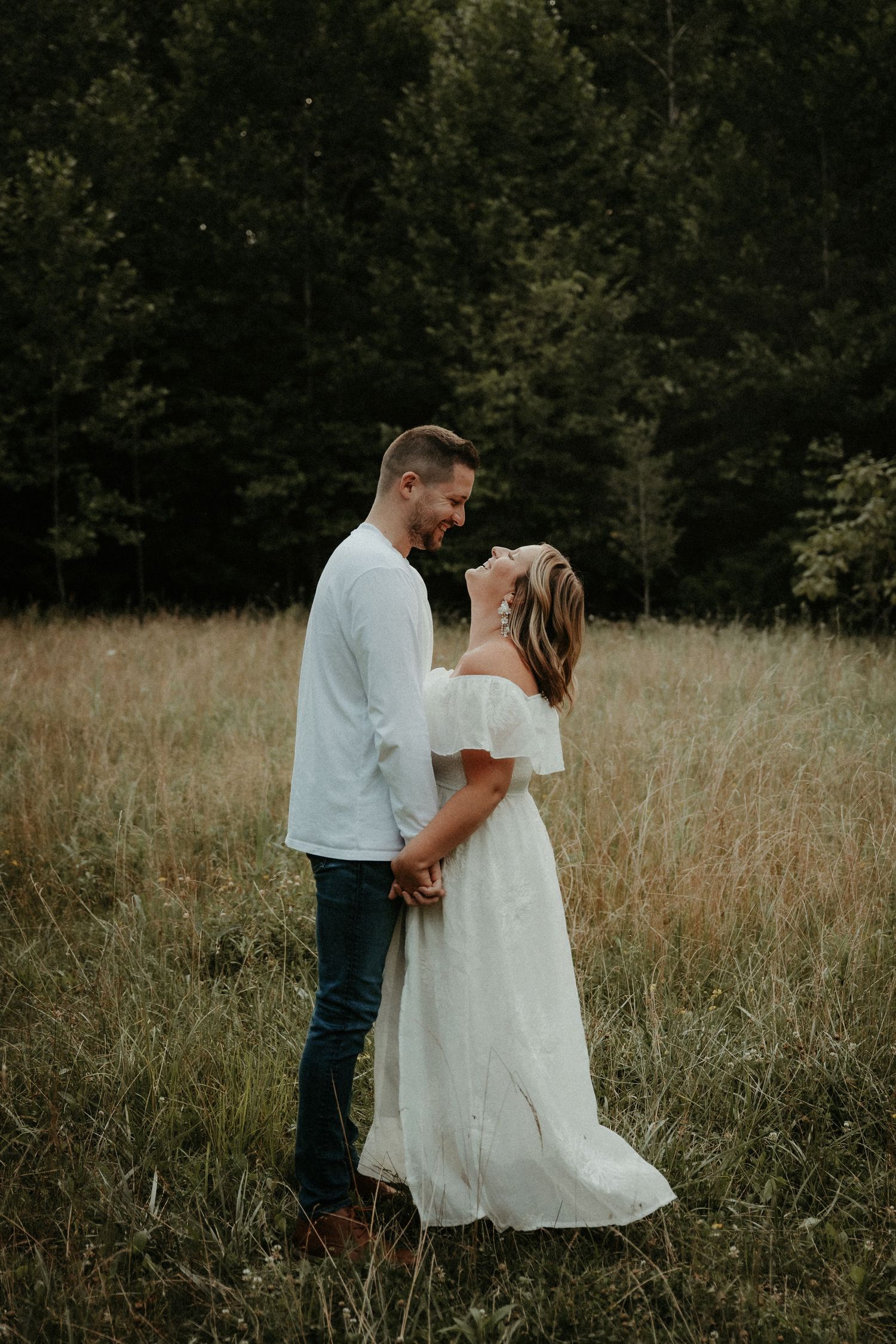 A romantic couple embraces in a tall grass field during a sunset photo session, wearing white dress and casual attire.