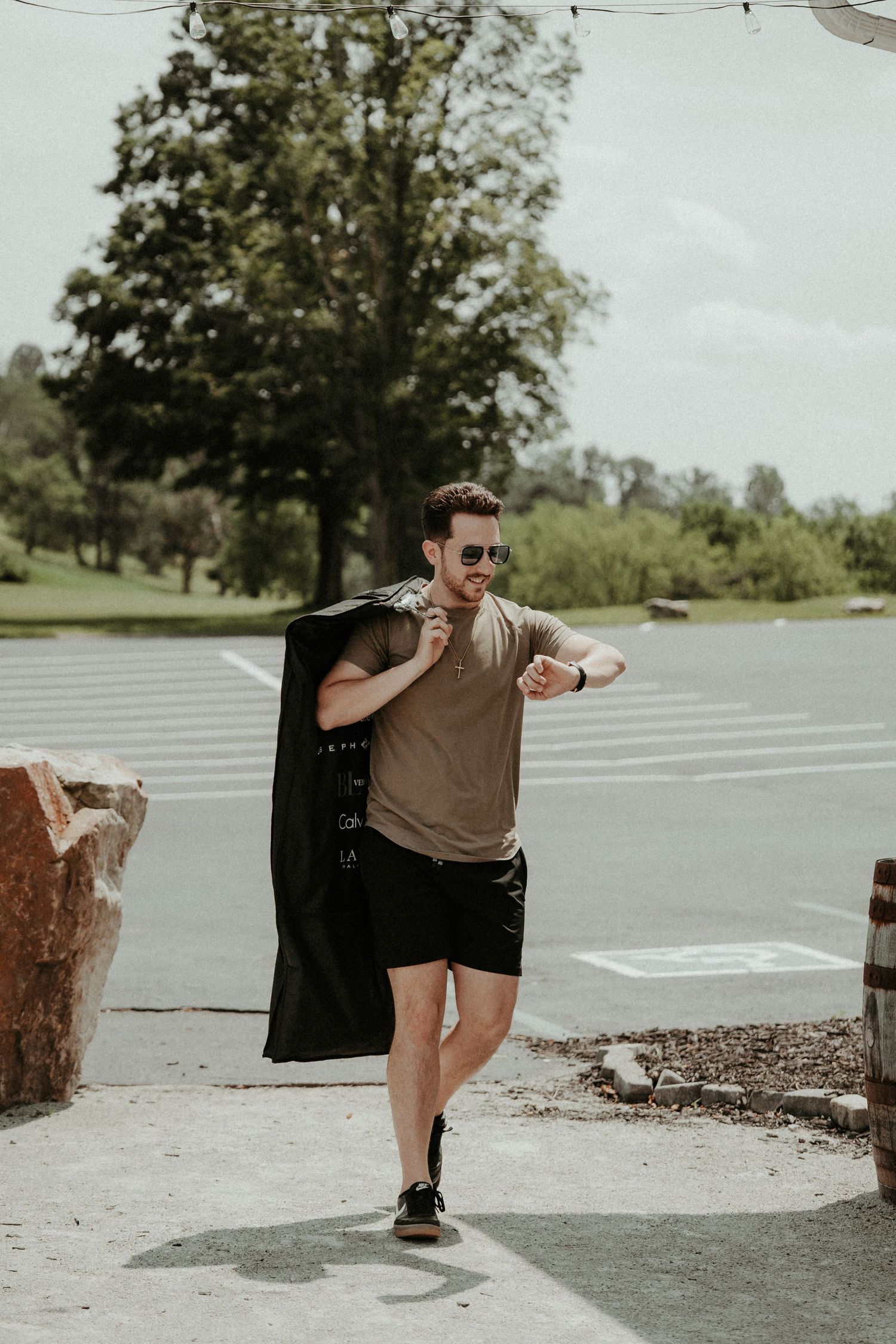 Person in casual attire checking watch while walking outdoors with jacket draped over shoulder on sunny day.