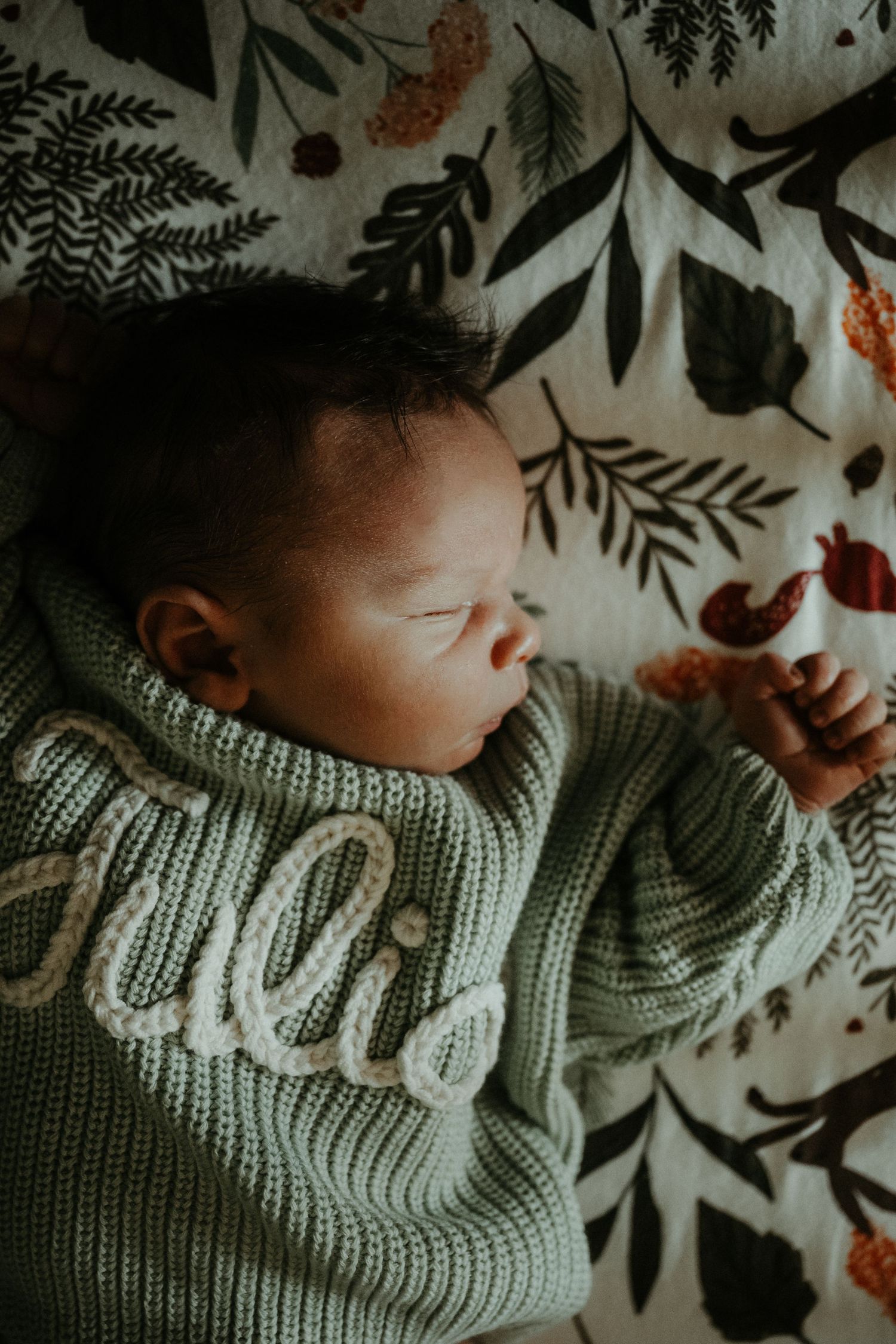 Sleeping baby wearing a gray knit sweater lies on floral print bedding in moody lighting. Pittsburgh / Westmoreland Photographer