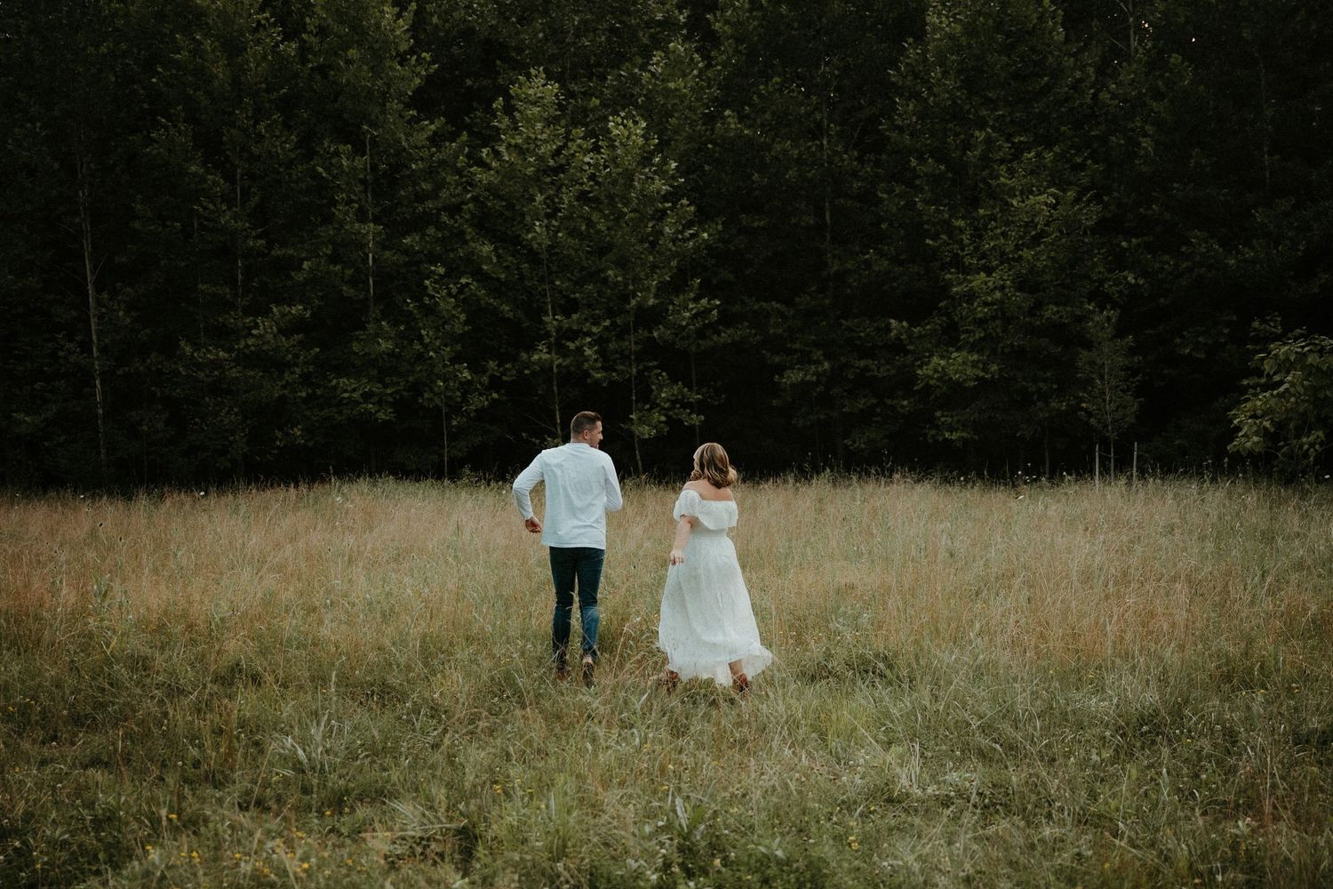 Two people in formal attire walk hand in hand through a golden meadow with a forest backdrop at dusk. Pittsburgh Wedding Photographer