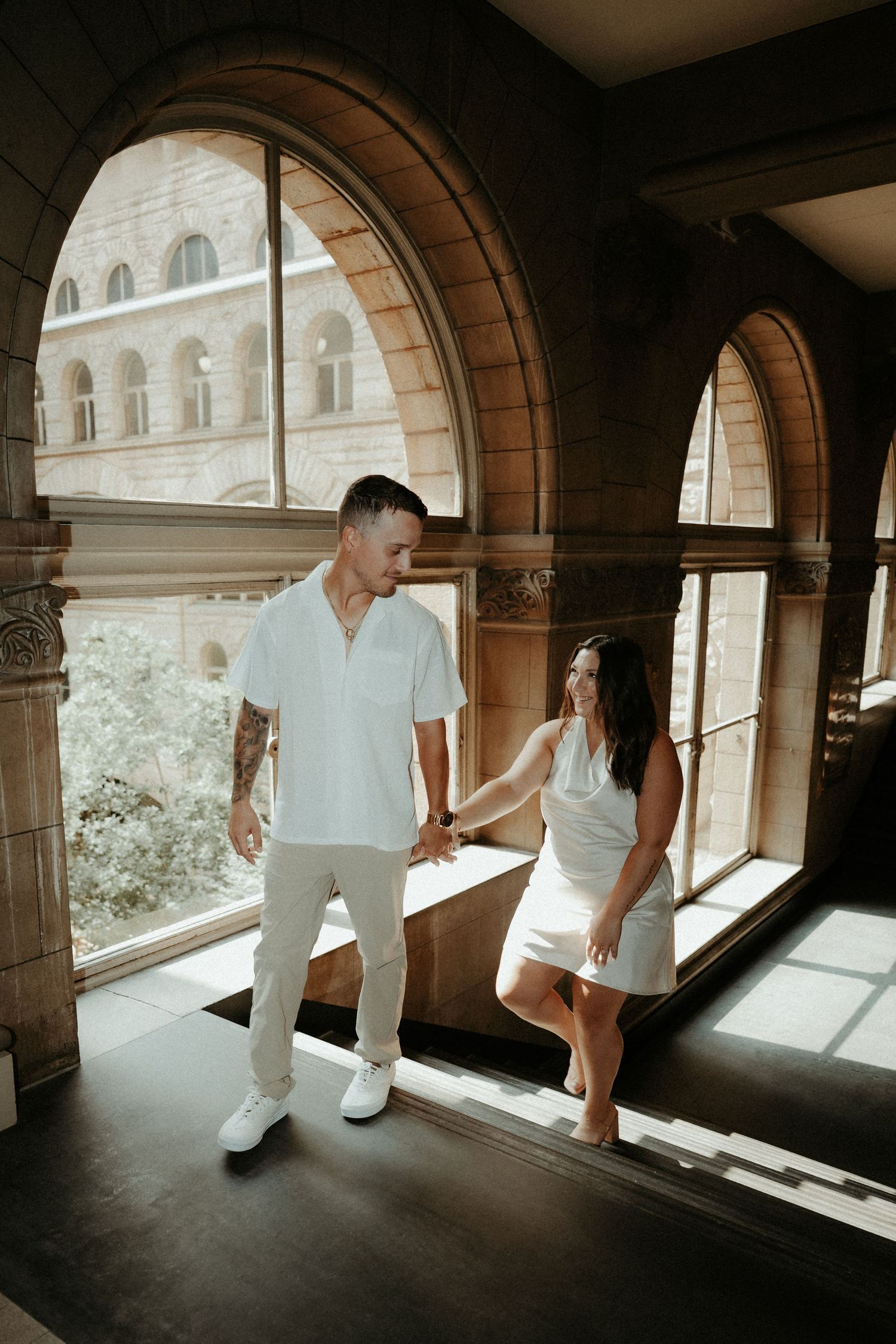 Couple in white outfits holding hands in an elegant arched hallway with architectural columns and natural lighting. Pittsburgh Wedding Photo