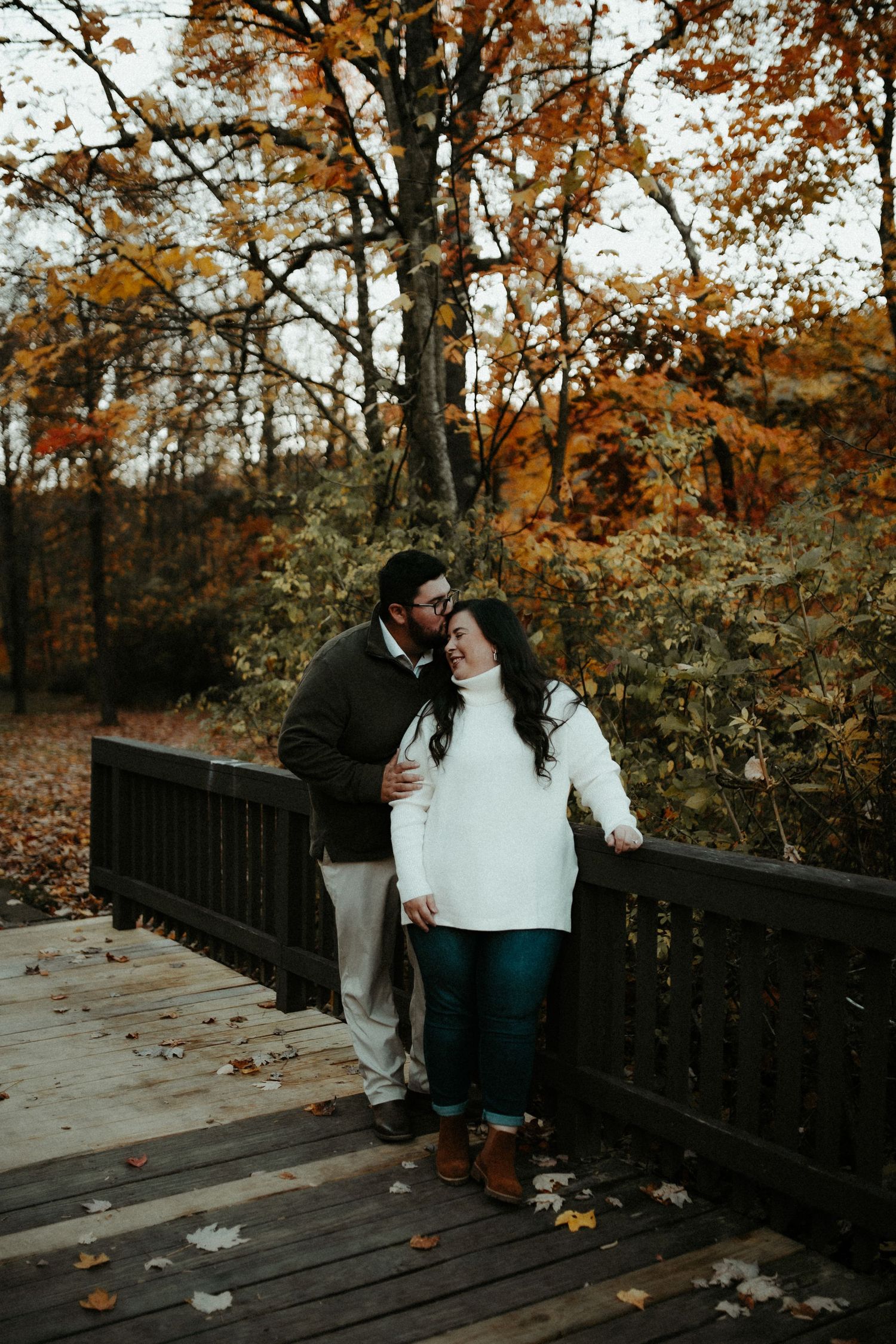Two people share an intimate moment on a wooden deck surrounded by autumn trees with golden leaves.