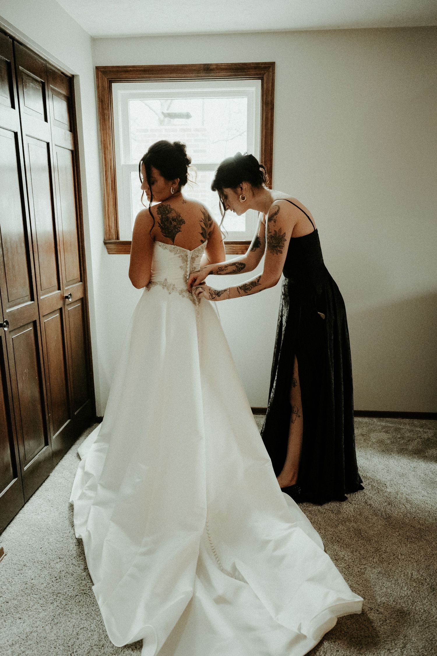 A bride in a flowing white dress gets help with her gown from someone in a black dress near a window. Pittsburgh Wedding Photographer