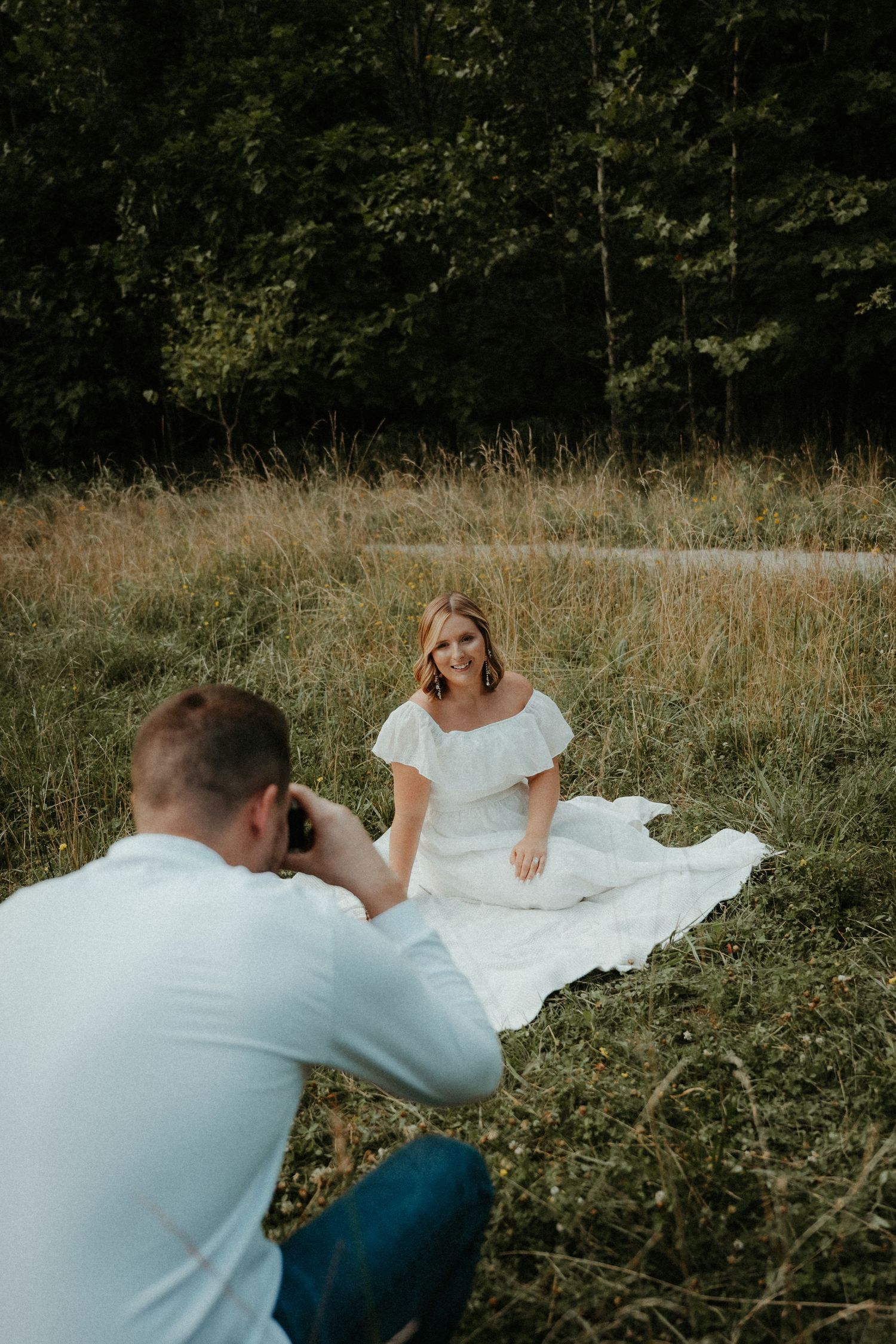 A man photographs his fiancé sitting on a blanket in a grassy meadow with trees in the background. Pittsburgh Wedding Photographer