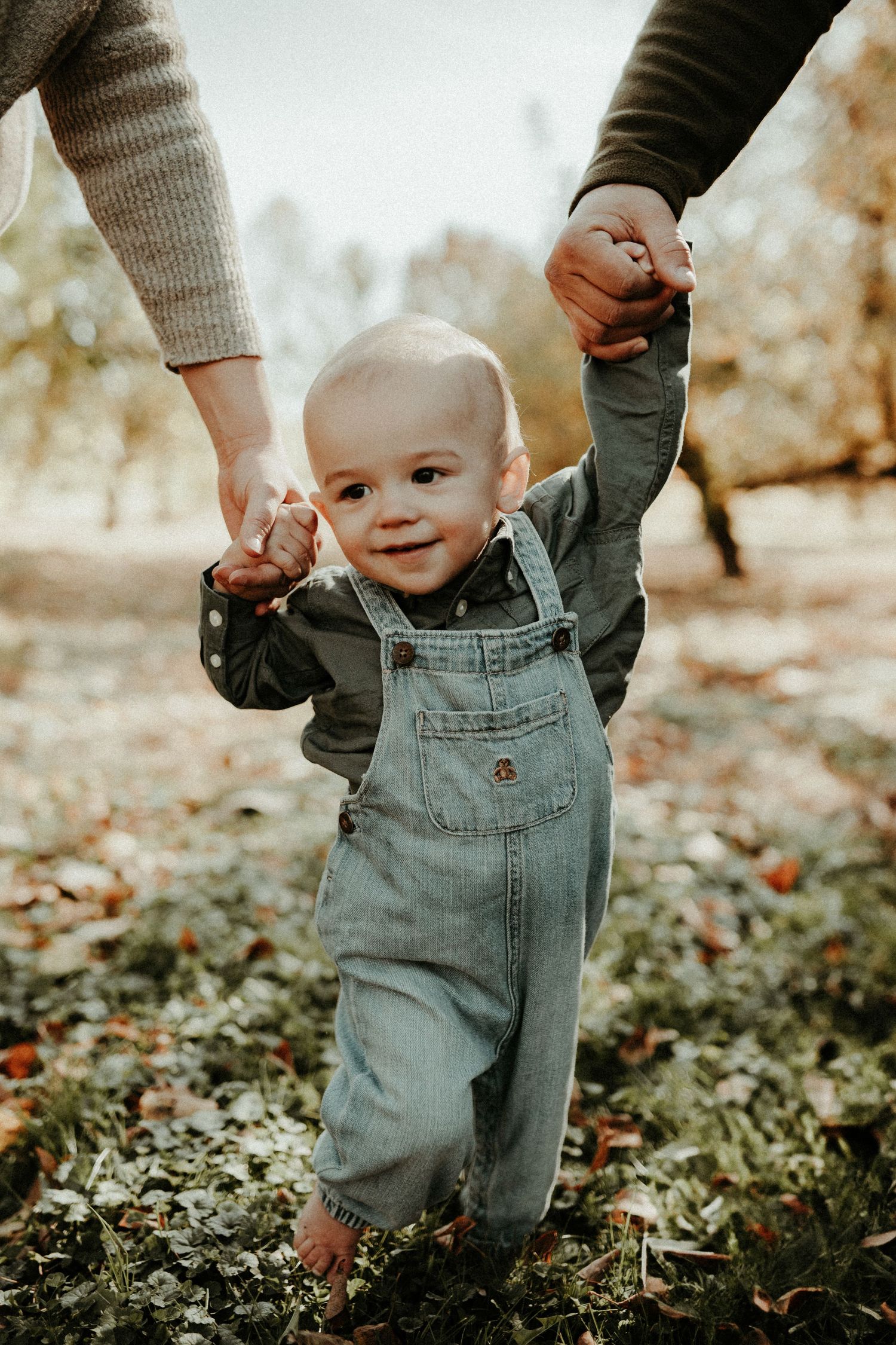 A smiling baby in denim overalls takes first steps while holding hands with adults. Pittsburgh Westmoreland Photographer