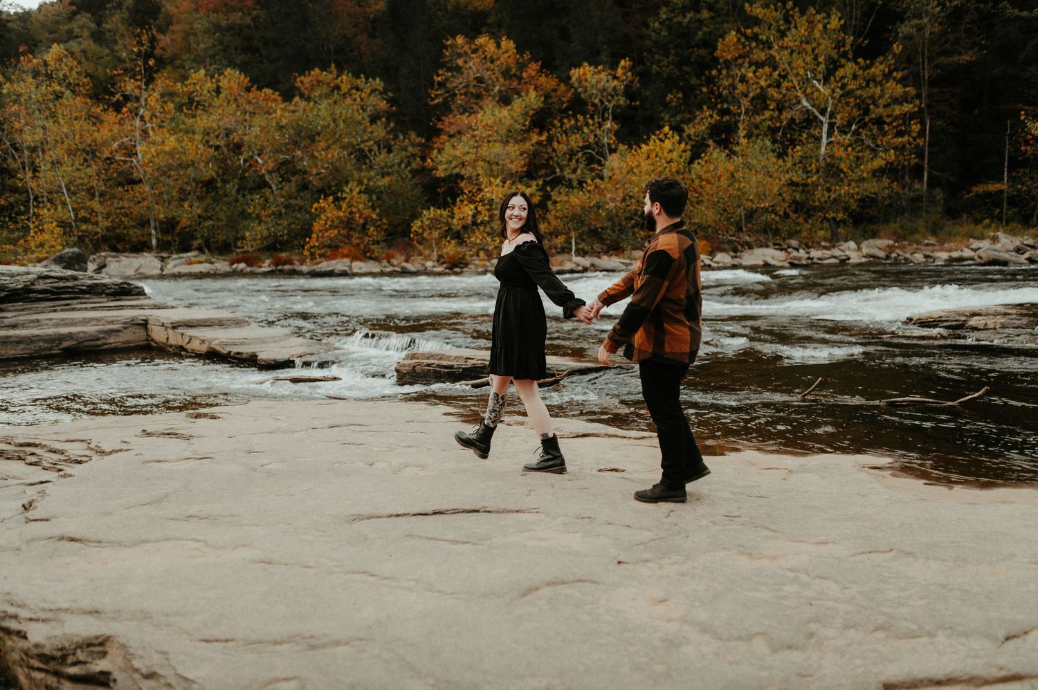 A couple walks across a rocky riverbed surrounded by autumn foliage during an outdoor photoshoot. Pittsburgh Wedding Photographer