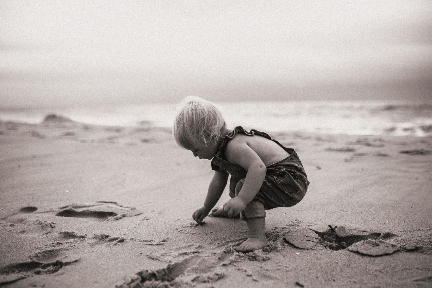 A child in overalls plays in the sand at the beach in black and white photography.