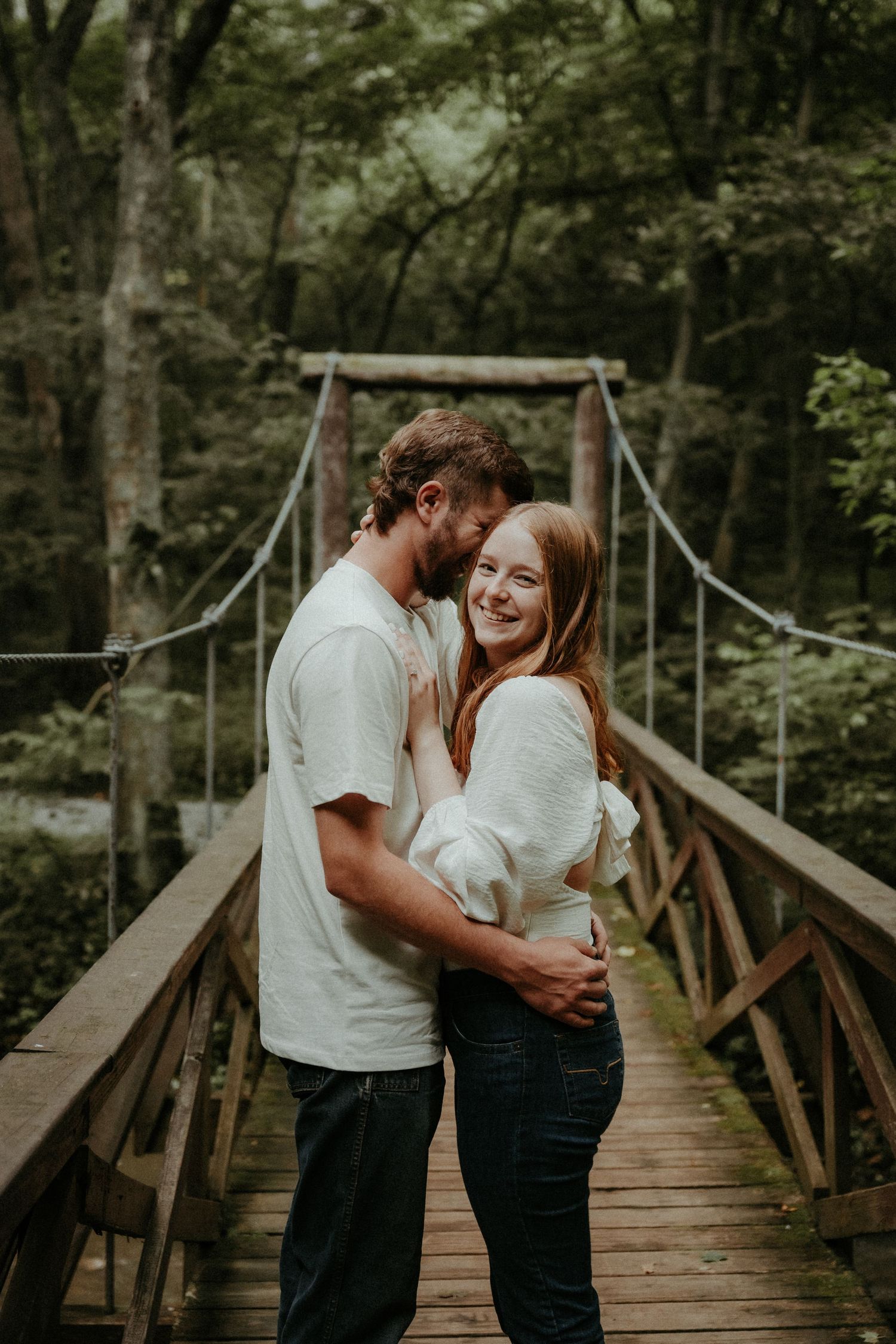 A couple shares intimate moments on a wooden footbridge in a lush forest setting during a romantic photoshoot.