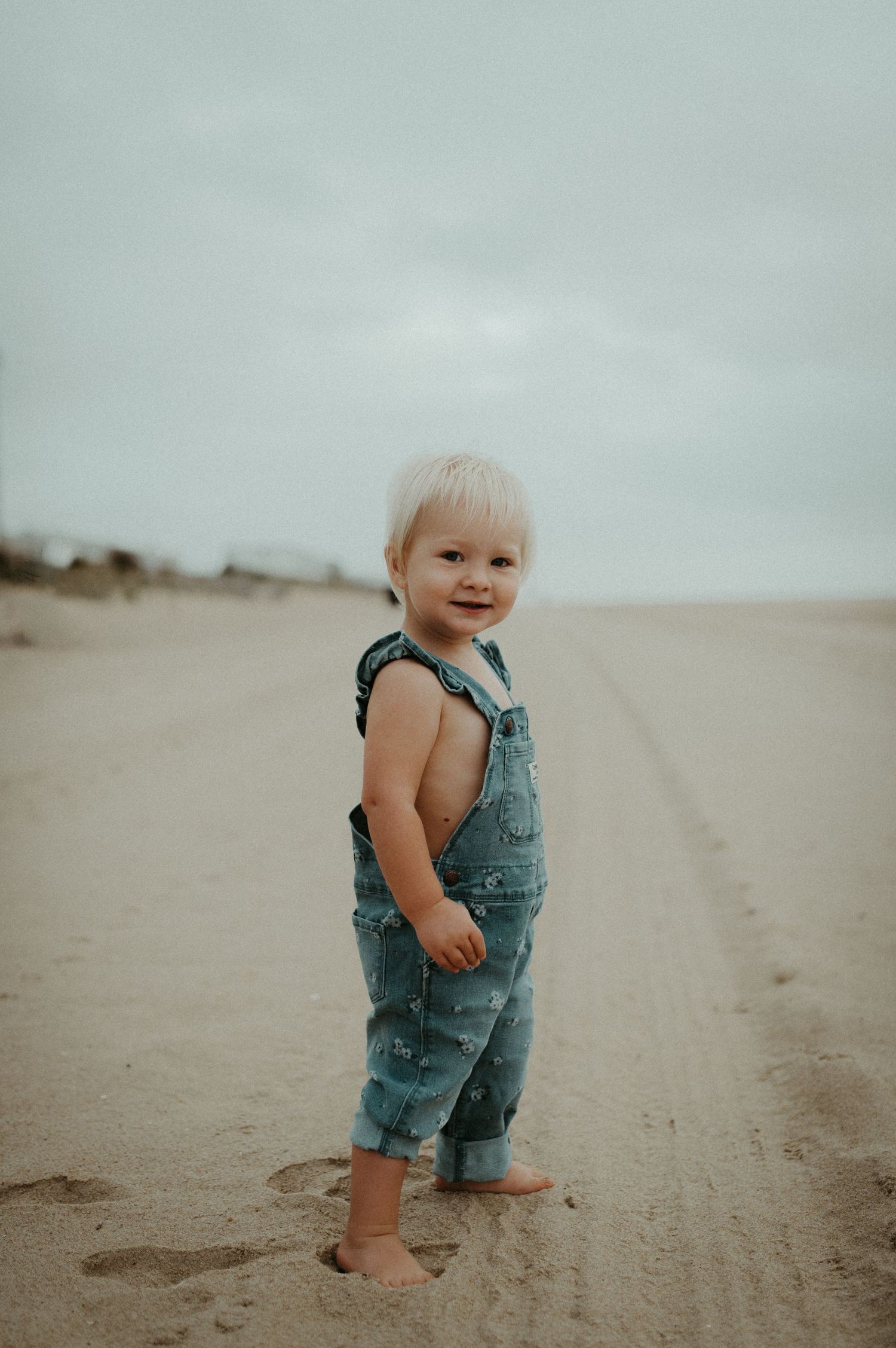 Toddler in denim overalls smiles while standing barefoot on a misty beach.
