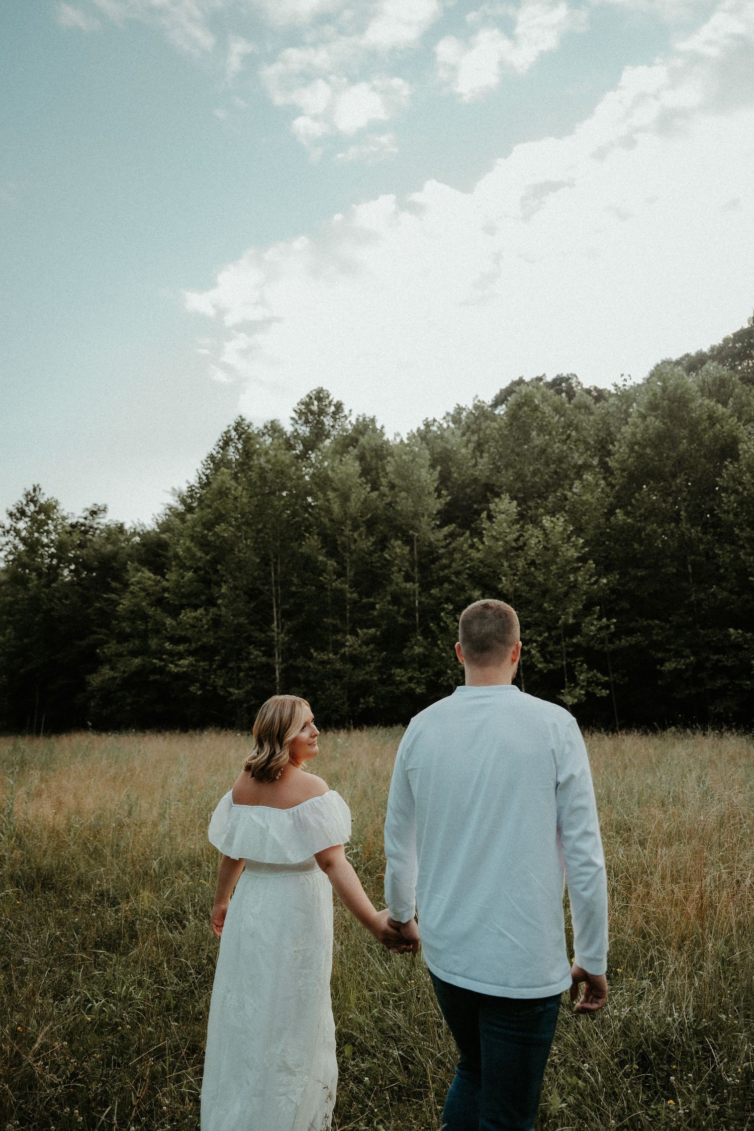 A couple walking hand in hand through a serene meadow with trees in the background on a cloudy summer day.