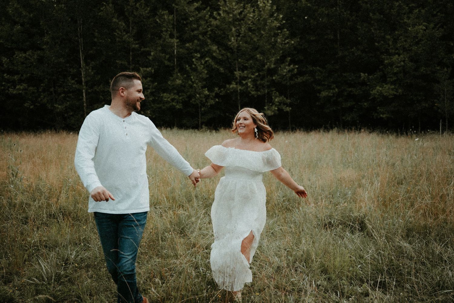 Happy couple runs playfully through a golden meadow at sunset with forest in the background. Pittsburgh Wedding Photographer