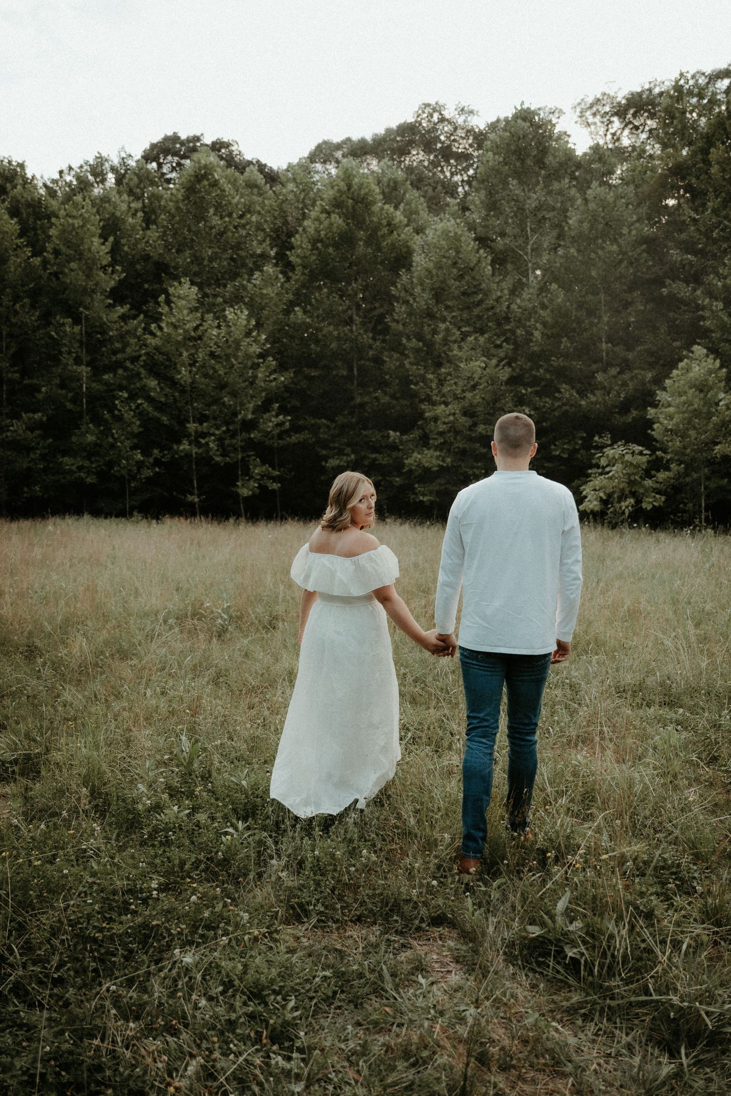 Couple walking through a peaceful wildflower meadow surrounded by trees at golden hour, holding hands in matching outfits.