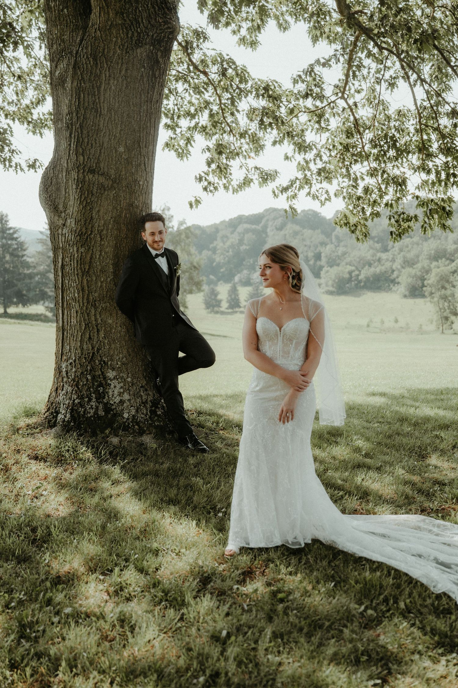 A wedding couple poses under a large tree in a scenic rural setting with rolling hills in the background. Pittsburgh Wedding Photographer