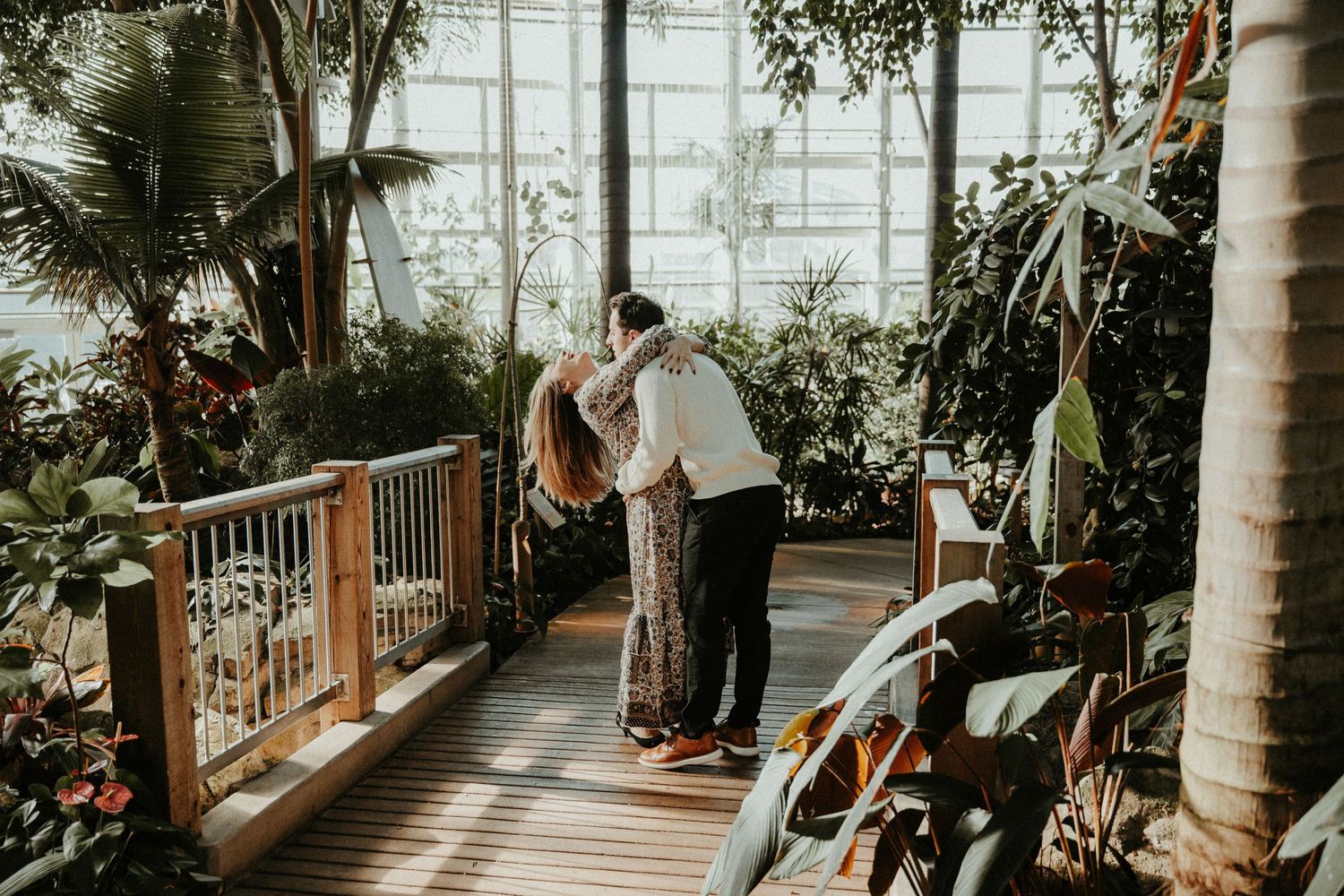 A couple shares an intimate moment on a wooden deck surrounded by lush greenhouse plants and natural light.
