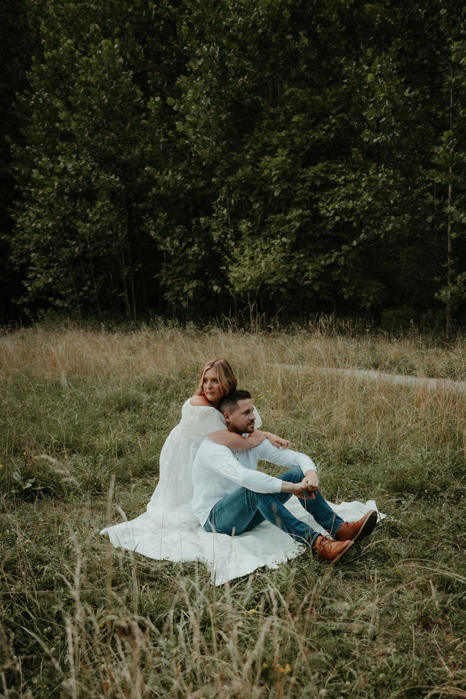 A couple sits together on a white blanket in a grassy meadow surrounded by trees on a romantic afternoon. Pittsburgh Wedding Photographer