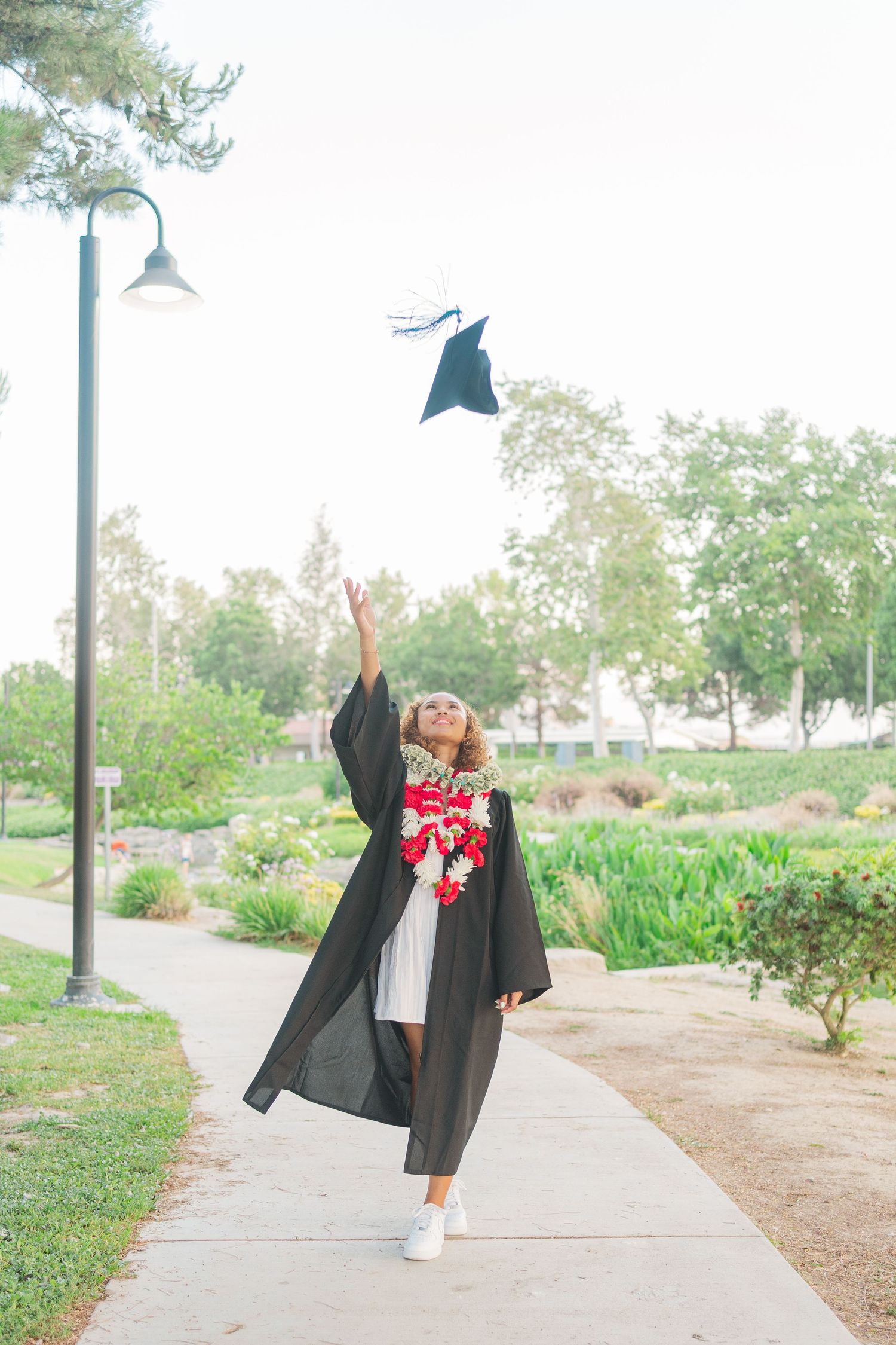 Graduate in black cap and gown throws mortarboard in air while celebrating on campus pathway surrounded by greenery.