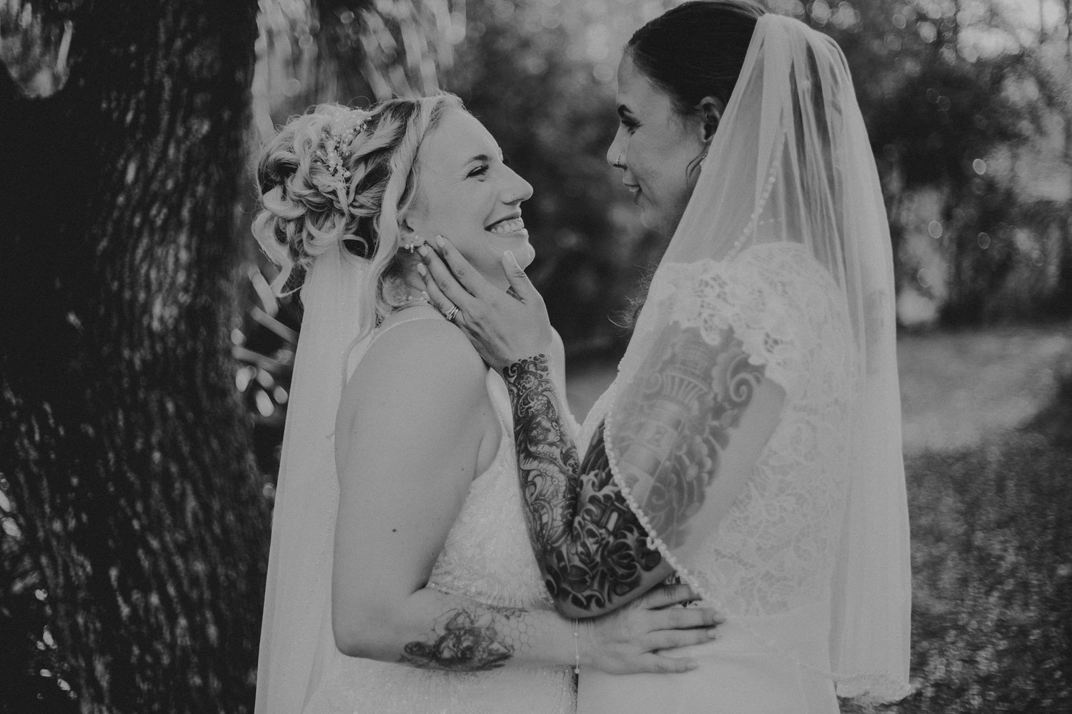 Two brides share a joyful moment together while wearing white wedding dresses and veils in black and white photography.