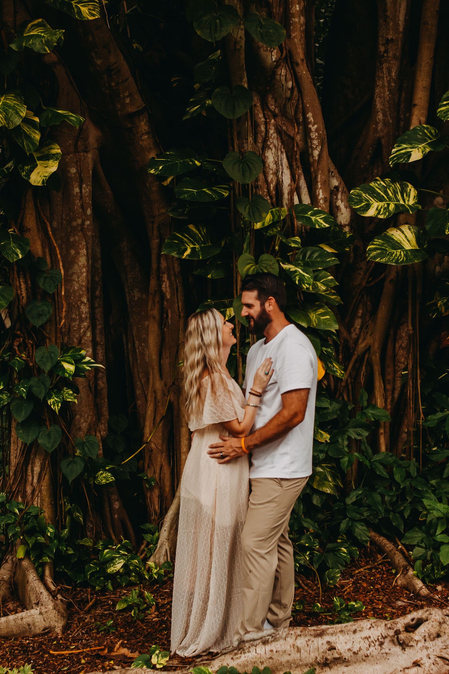 A couple shares an intimate moment among tall trees and lush greenery in a romantic outdoor setting.