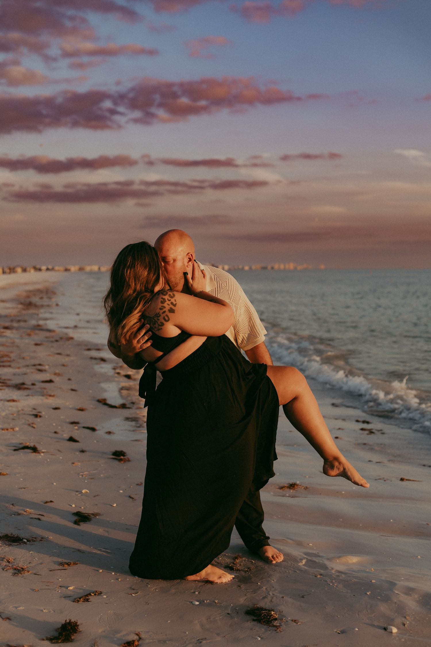 A couple embraces on a sandy beach at sunset with pink clouds and ocean waves in the background.