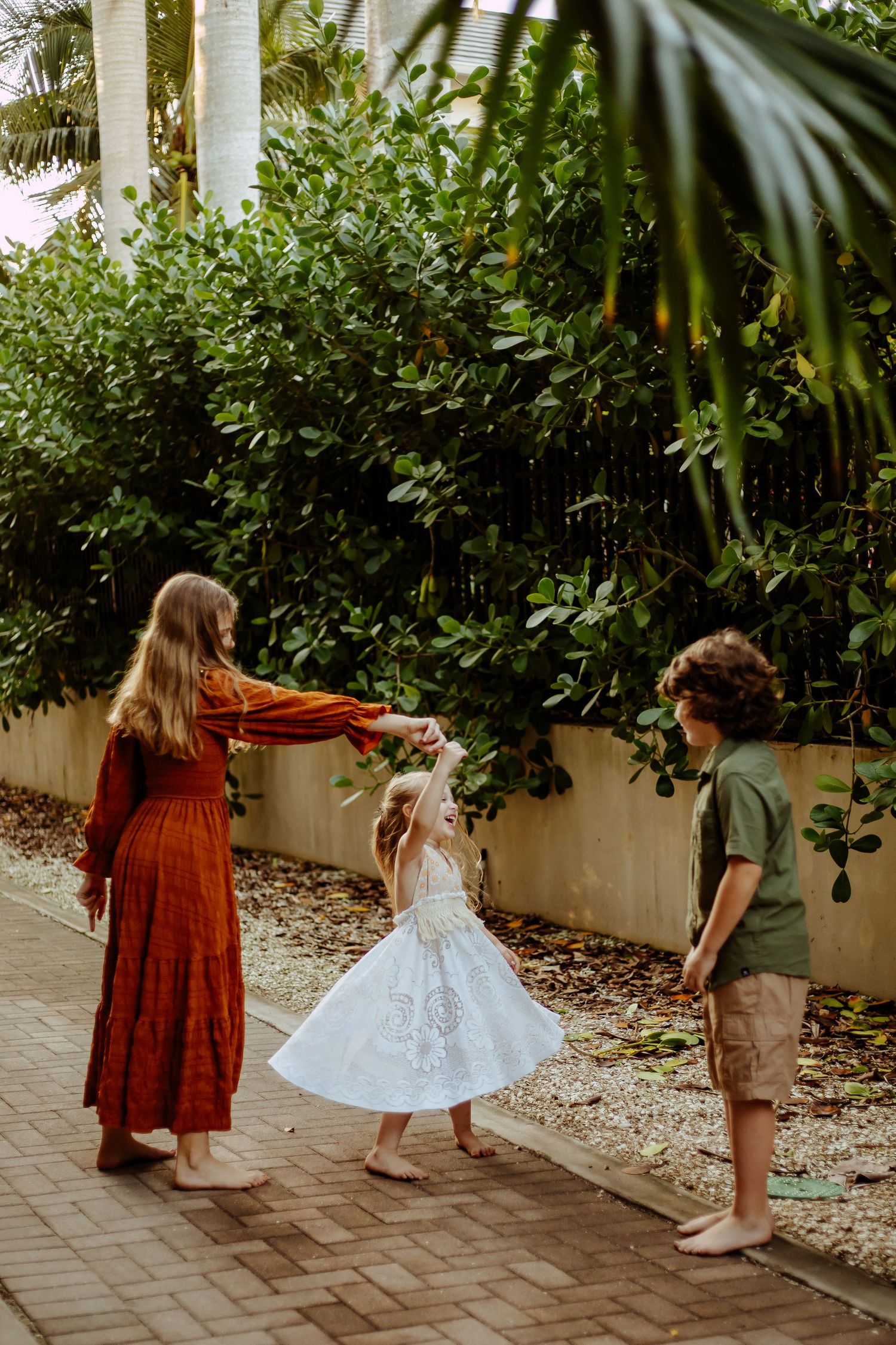 Three people play together on a brick path surrounded by lush tropical garden foliage and palm trees.