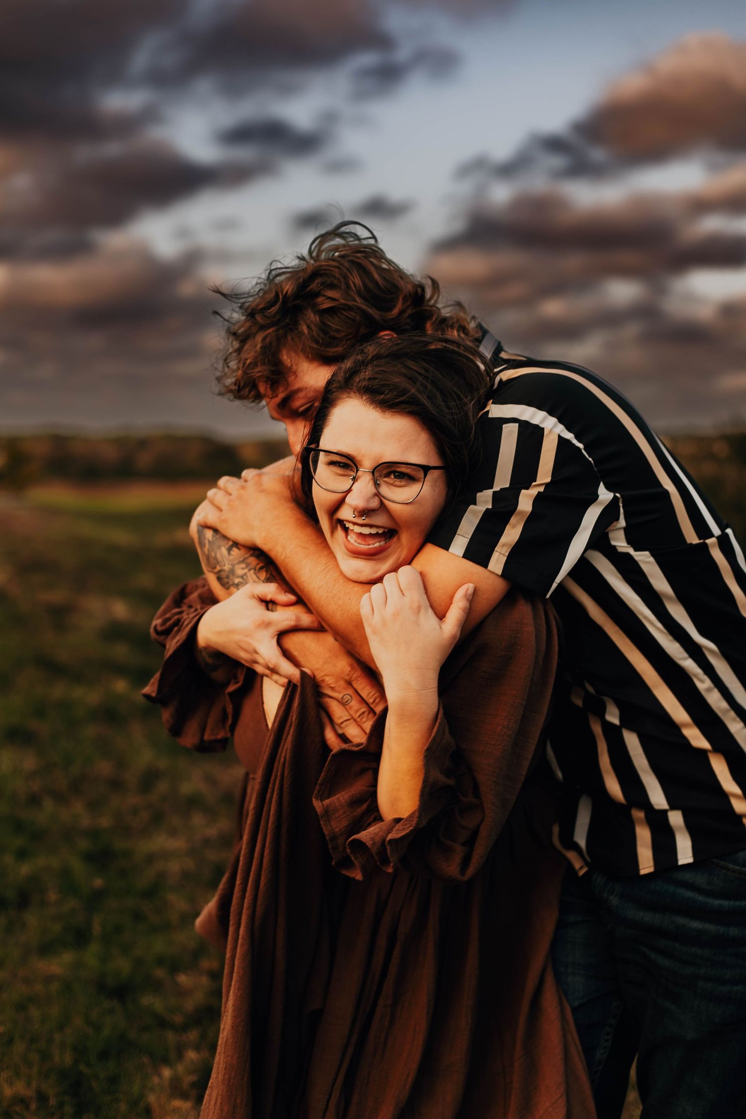 A person in glasses and a striped shirt laughs joyfully while posing outdoors at sunset.