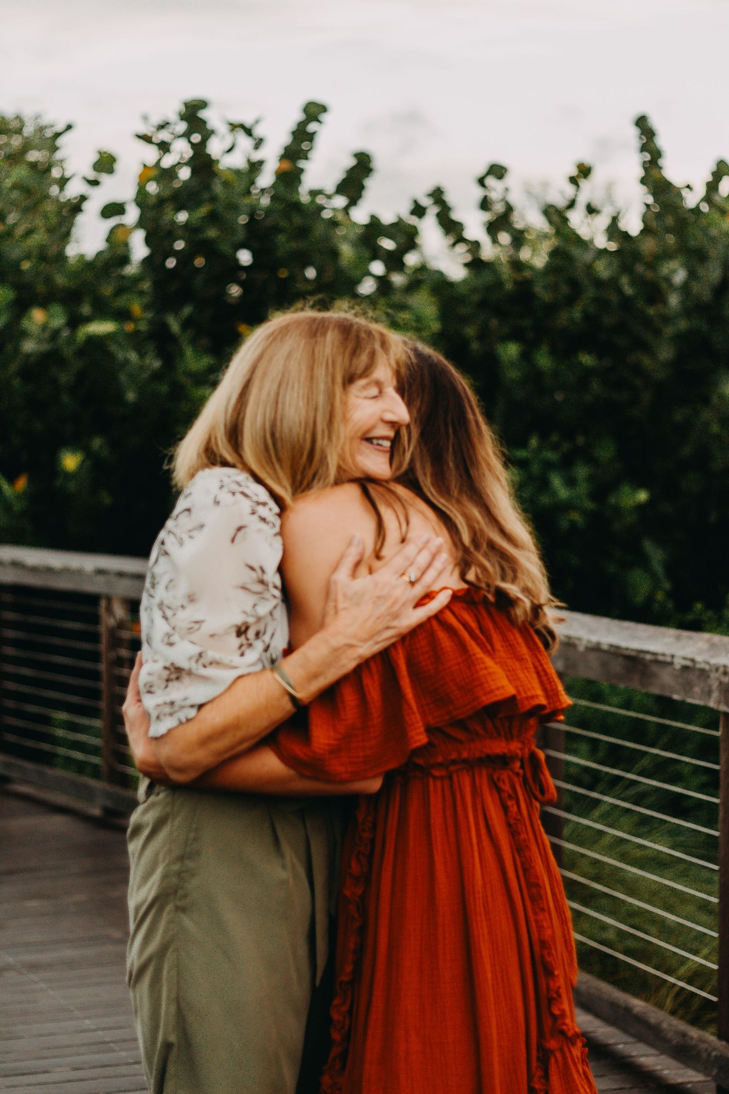 Two people share a tender embrace outdoors, one wearing a rust-colored dress and the other in olive green.