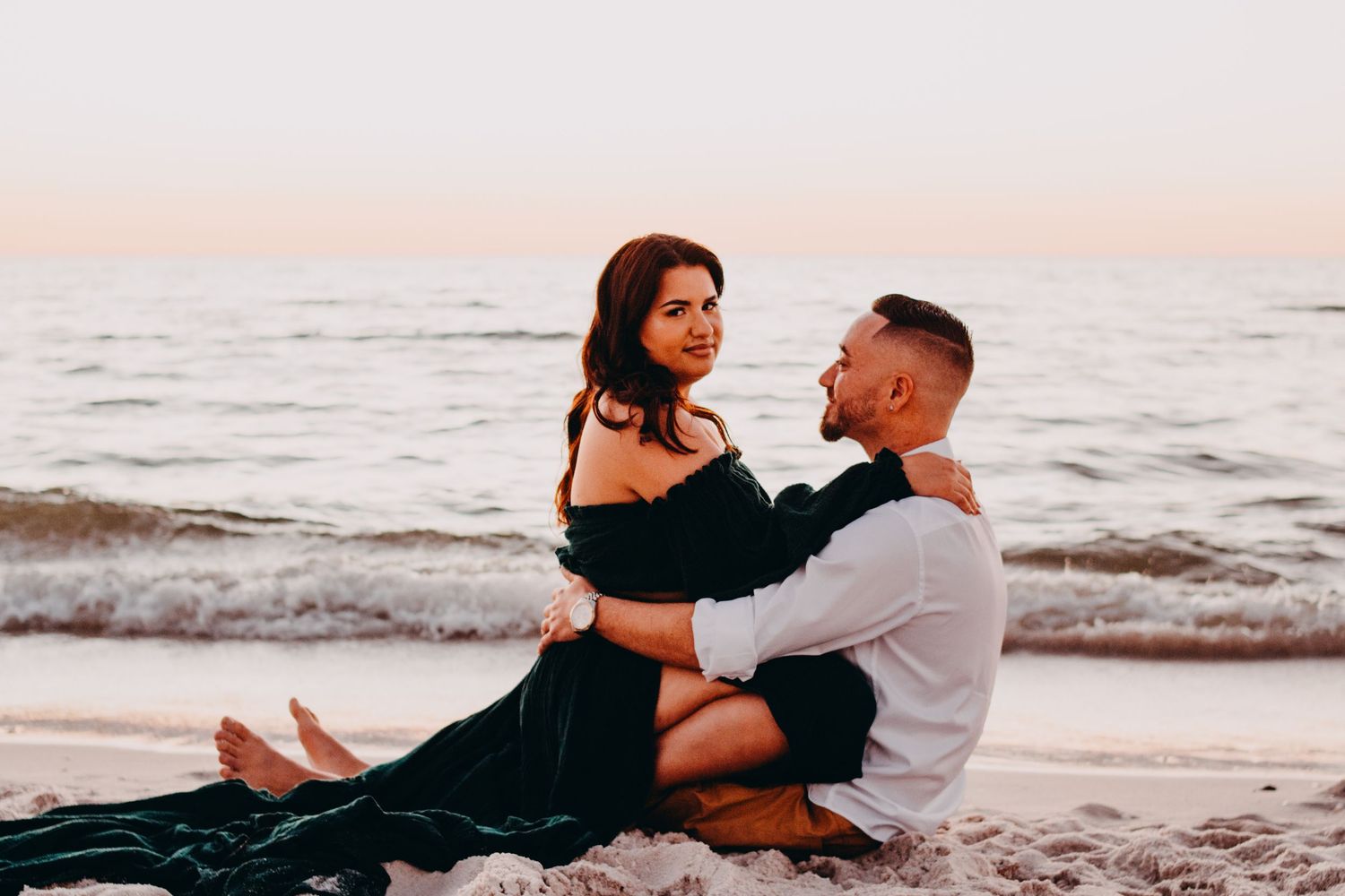 A romantic couple embraces on a sandy beach at sunset with ocean waves in the background.