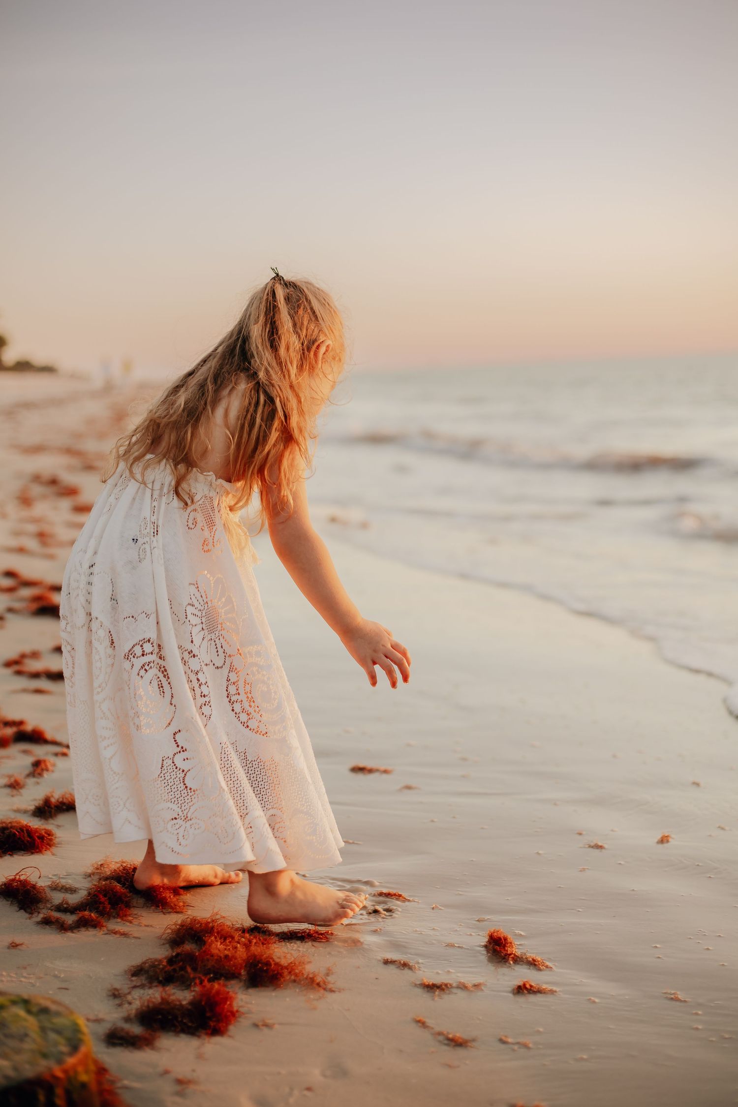A figure in a white lace dress bends down to touch the ocean waves at sunset on a peaceful beach.