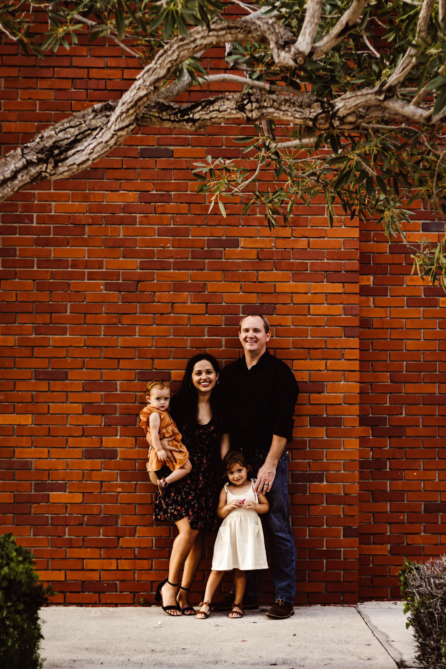 A family sits together against a red brick wall outdoors with tree branches visible overhead.