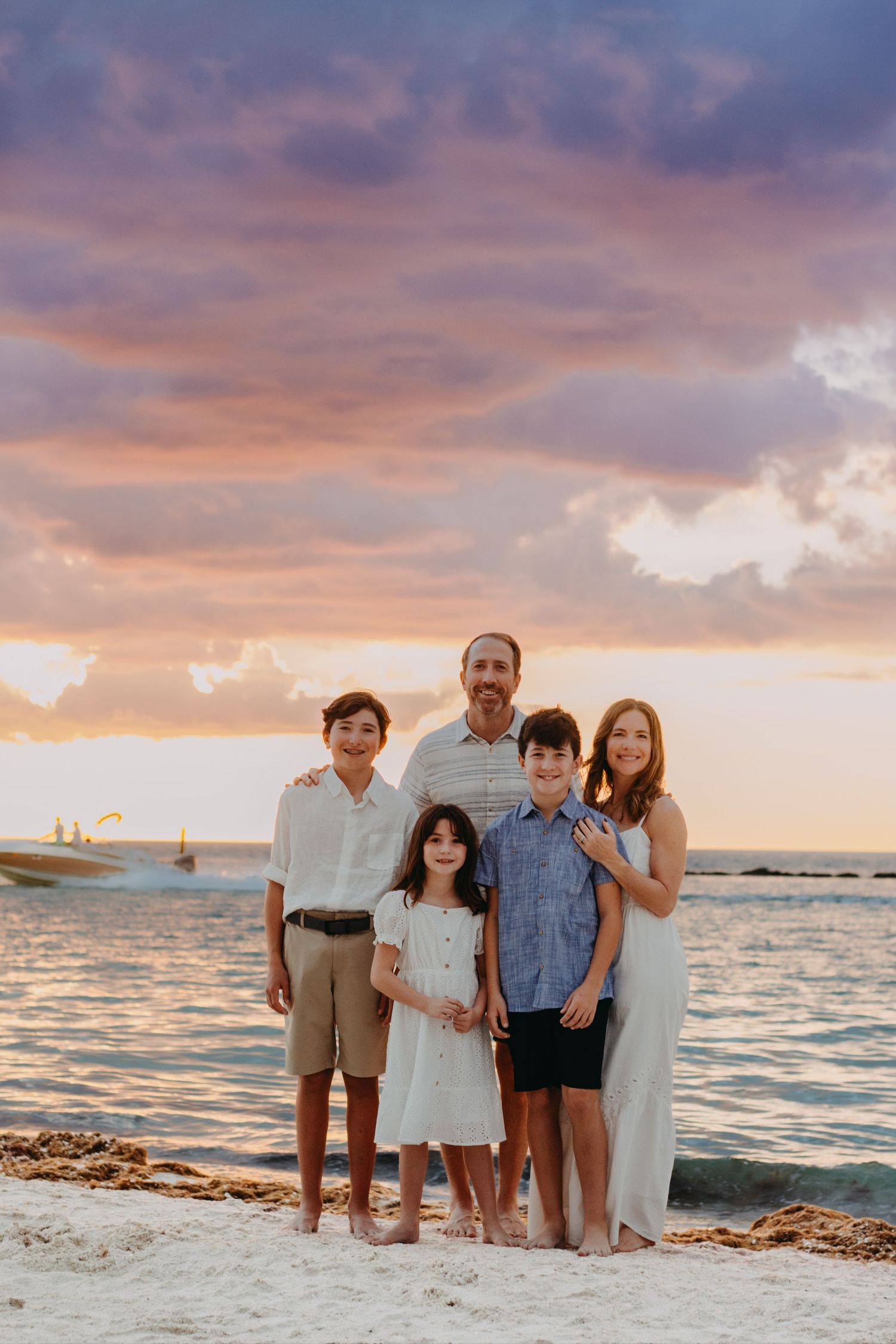 A family stands together on a beach at sunset with golden clouds and ocean waves in the background.