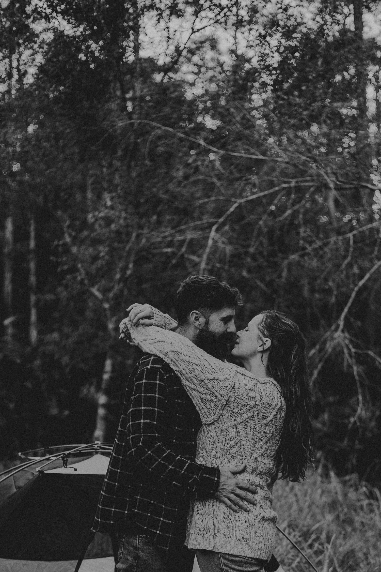 A romantic black and white photo of a couple embracing in a forest setting with a guitar case visible.