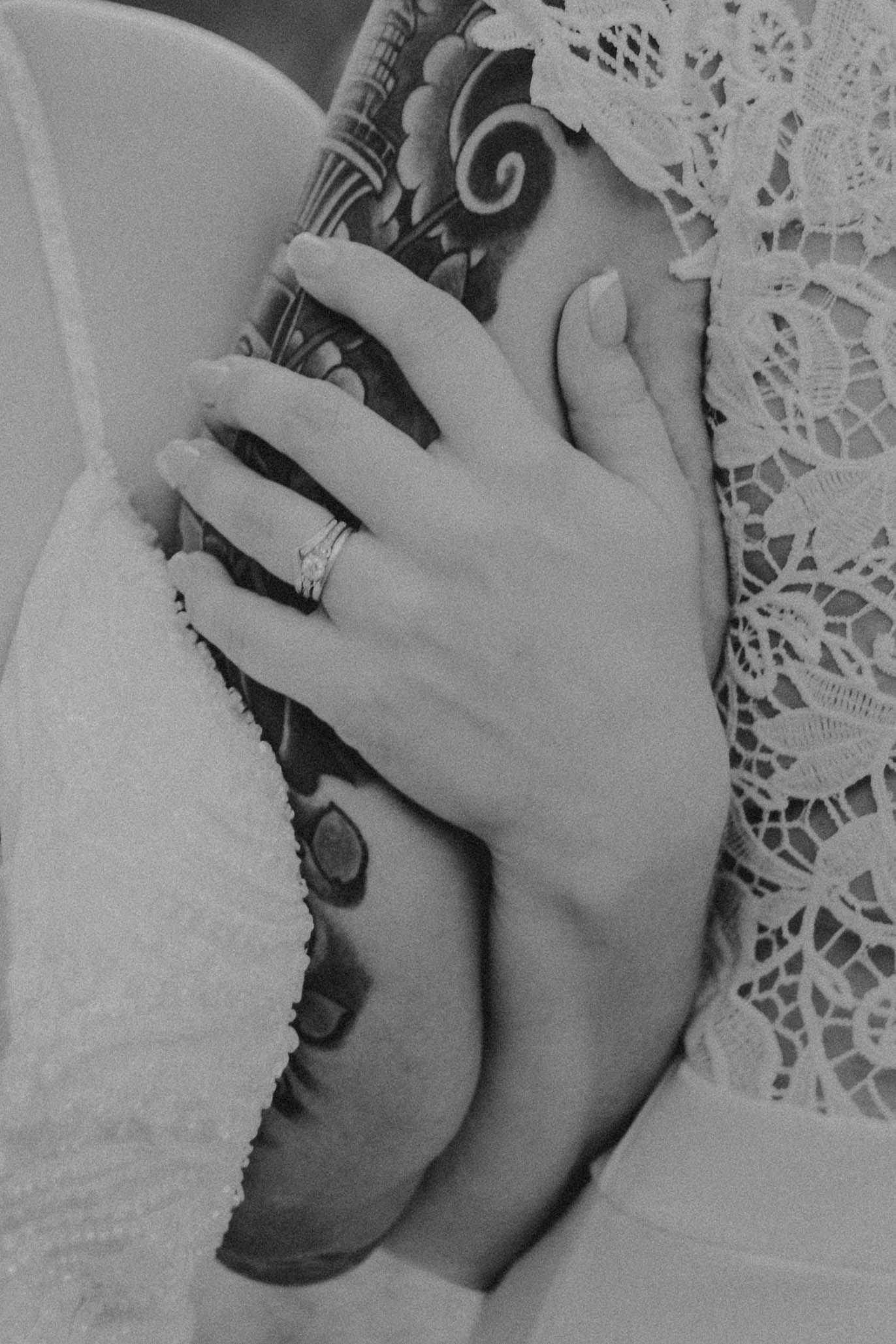 A delicate hand wearing a ring rests against white lace fabric and dark tattoo artwork in black and white.