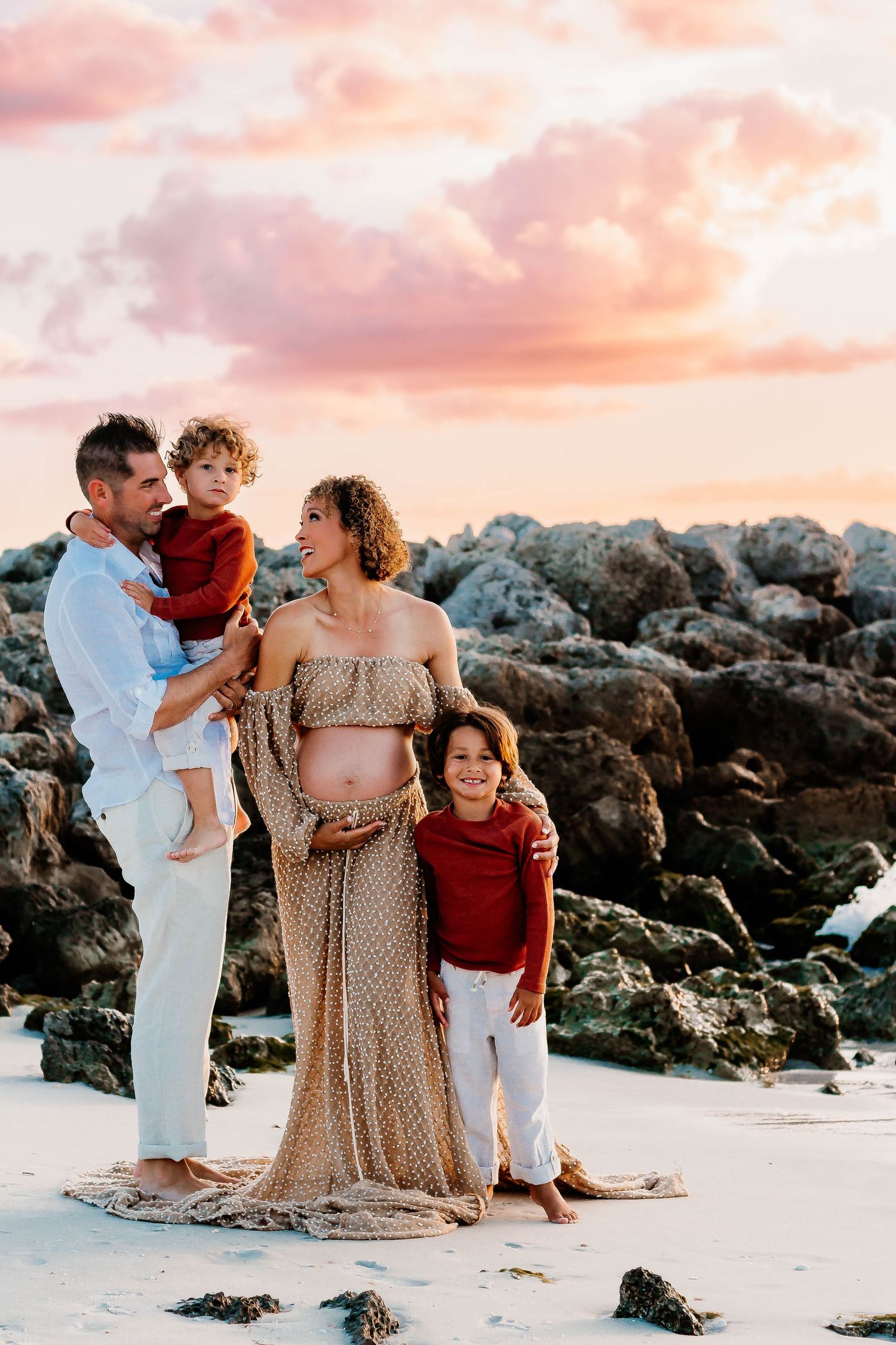 Family enjoying a sunset moment together on a rocky beach with pink and orange sky in the background.