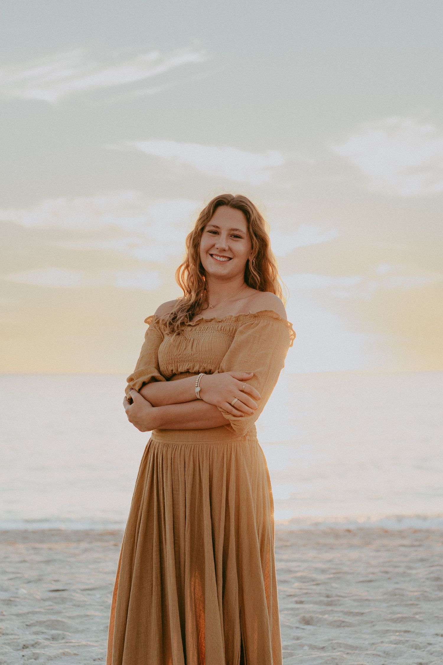 Woman in a flowing caramel-colored dress poses by the ocean at sunset.