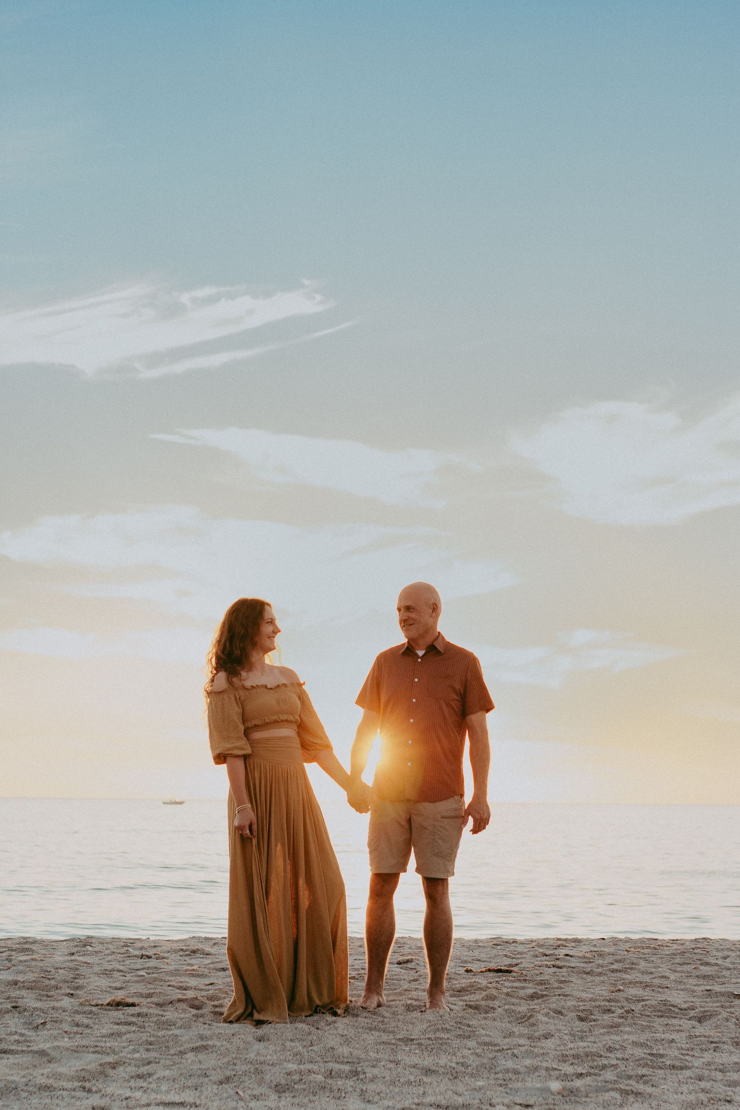 A couple stands together on a sandy beach at sunset with warm golden light glowing behind them.
