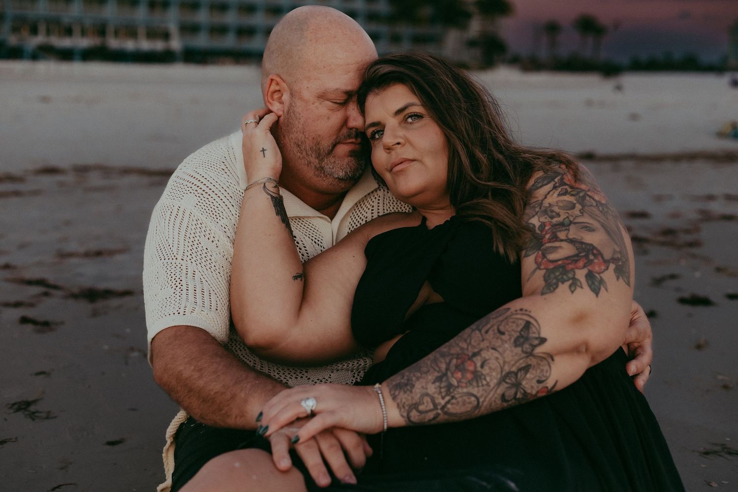 Intimate couple embraces on beach at sunset, showing tattoos and wearing black dress and white sweater.