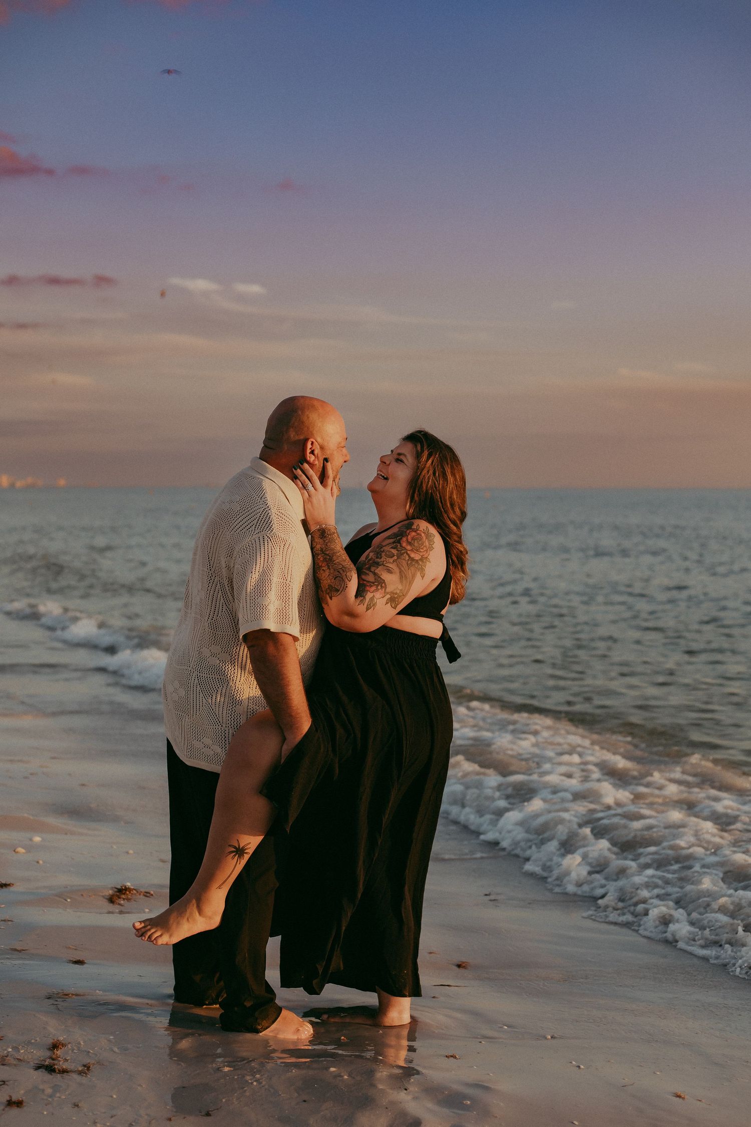 A couple shares an intimate moment sitting together on a sandy beach at sunset with ocean waves in the background.