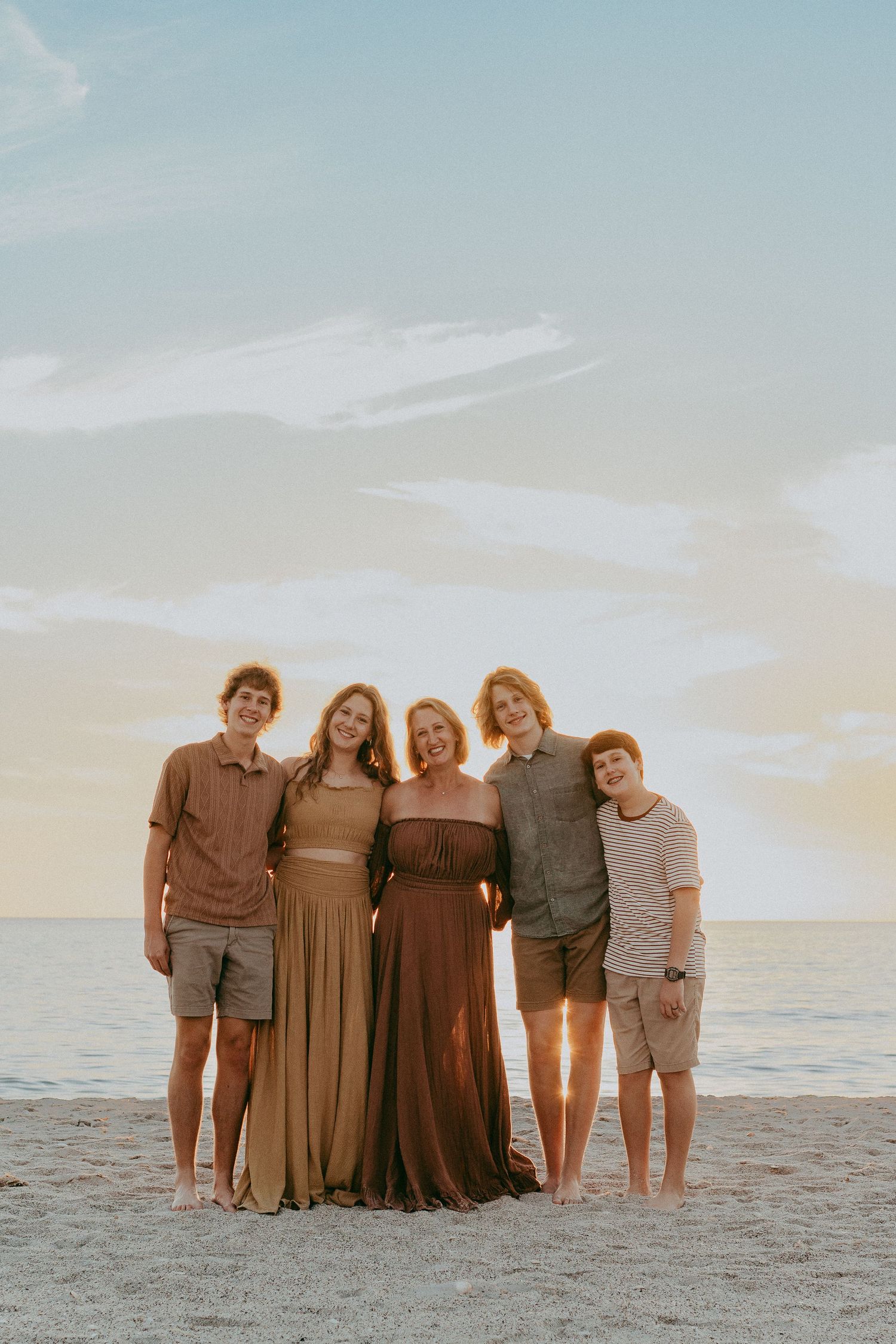 A group in coordinating earth-tone outfits poses together on a sandy beach at sunset.