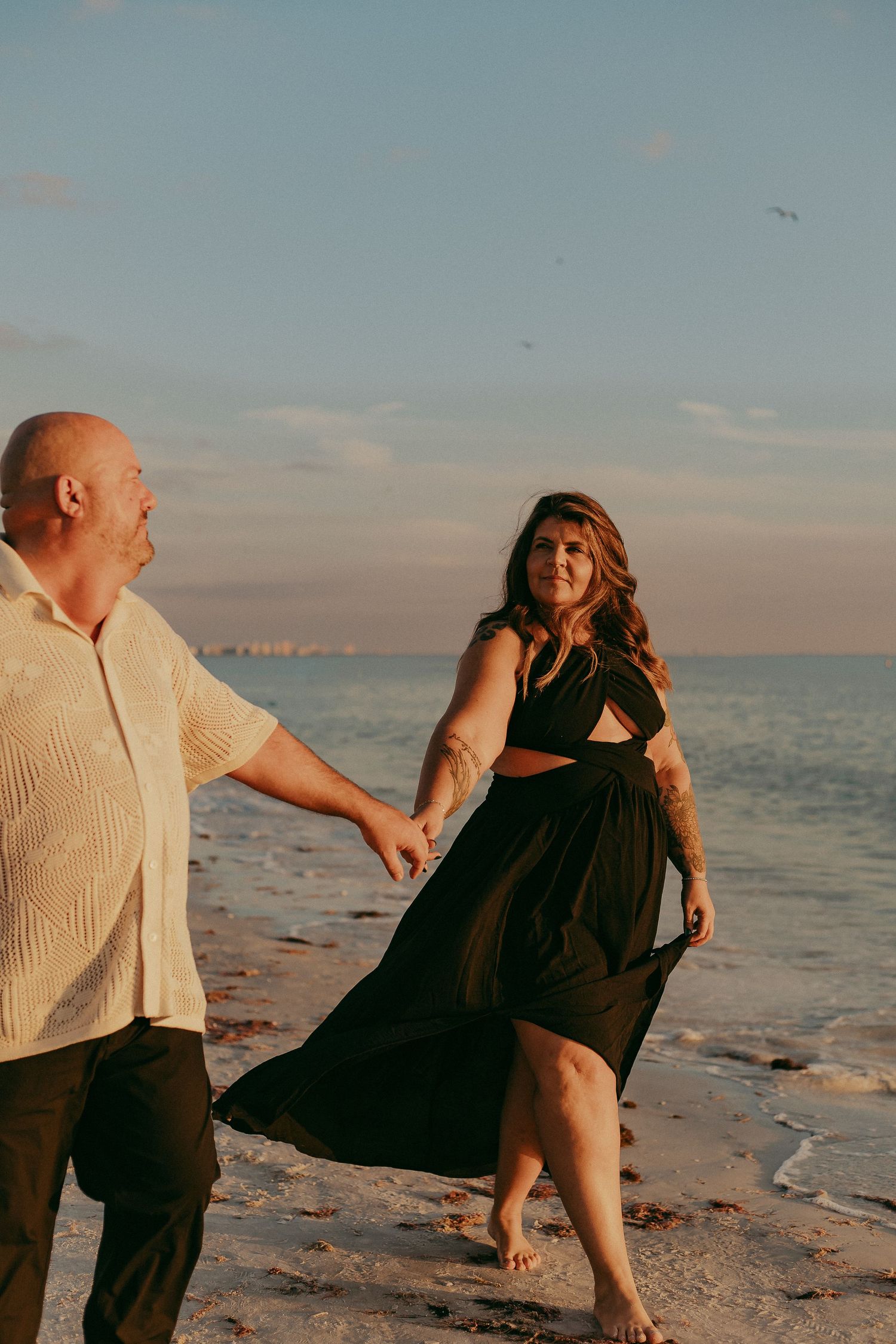 A couple holds hands while walking along a beach at sunset, with the woman wearing a flowing black dress.