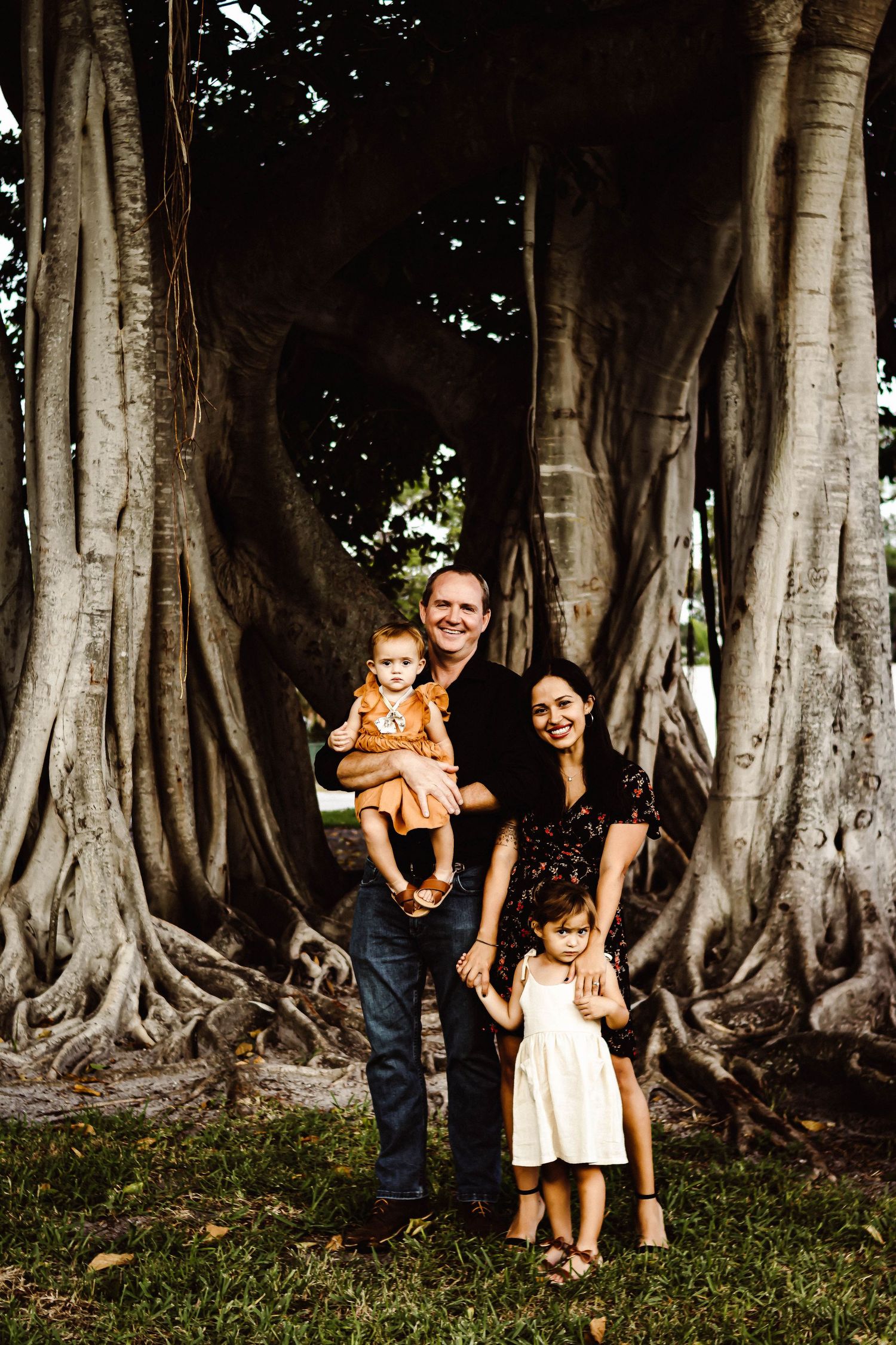 A family poses together near large banyan trees with dramatic aerial roots in a park setting.