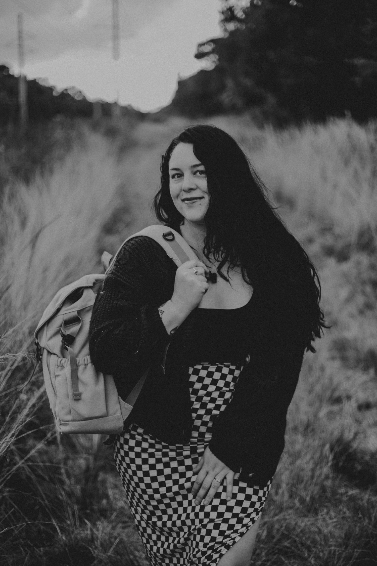 Black and white portrait of someone in a checkered dress and dark cardigan holding a backpack in a grassy outdoor setting.