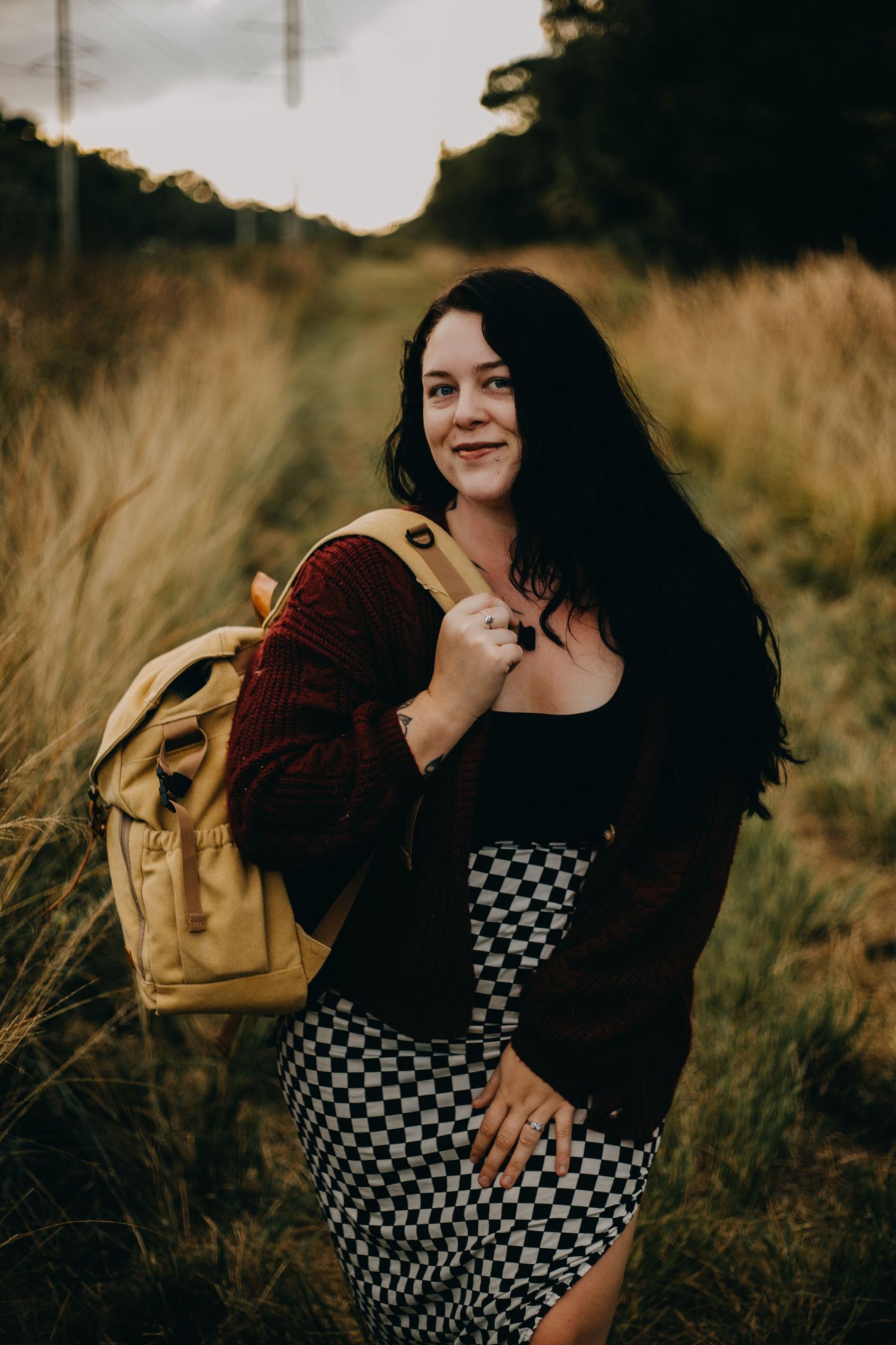 A couple embraces in tall grass at sunset wearing a checkered dress and burgundy sweater with a tan backpack.