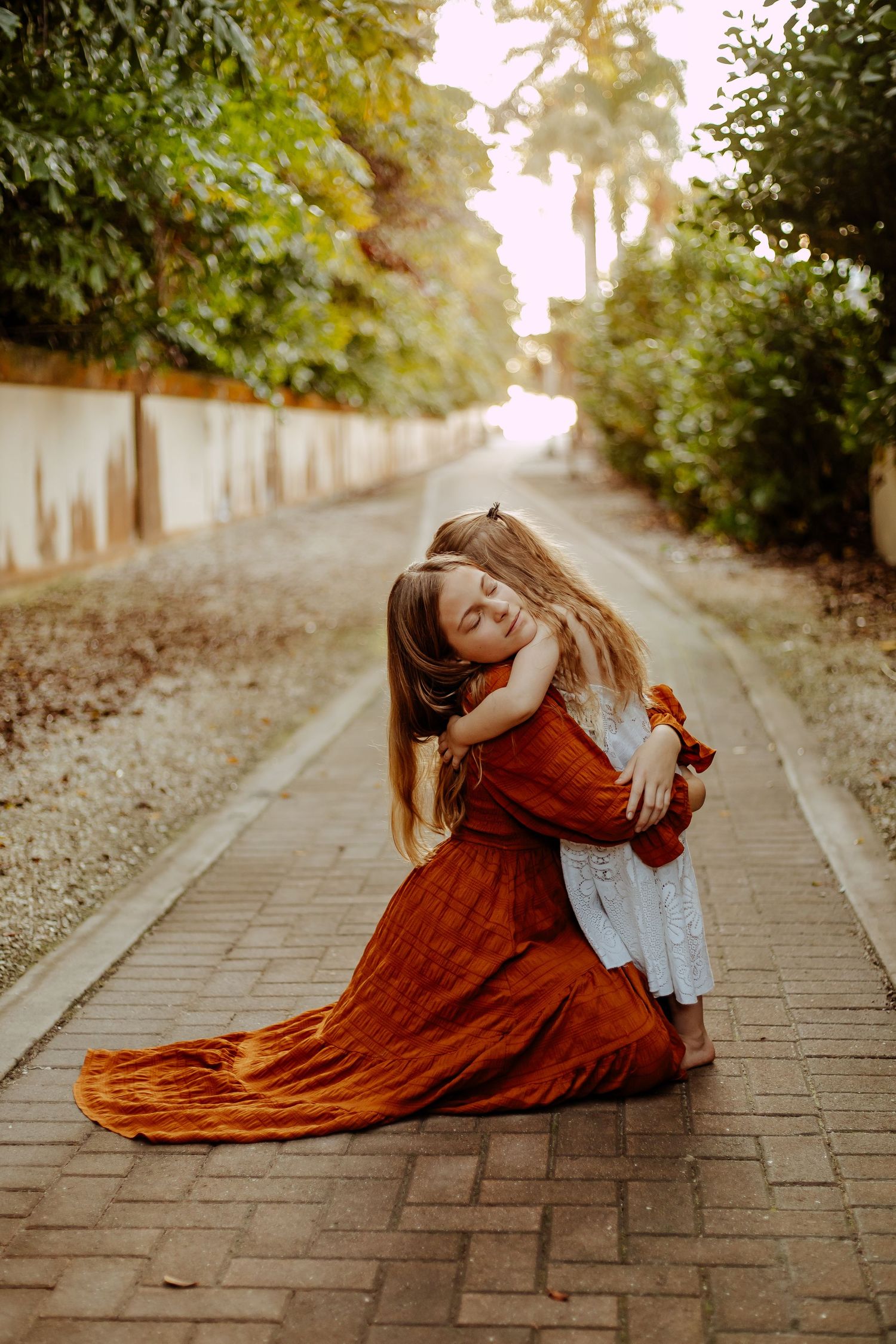A person in a flowing rust-colored dress lies gracefully on a brick pathway bordered by green trees.