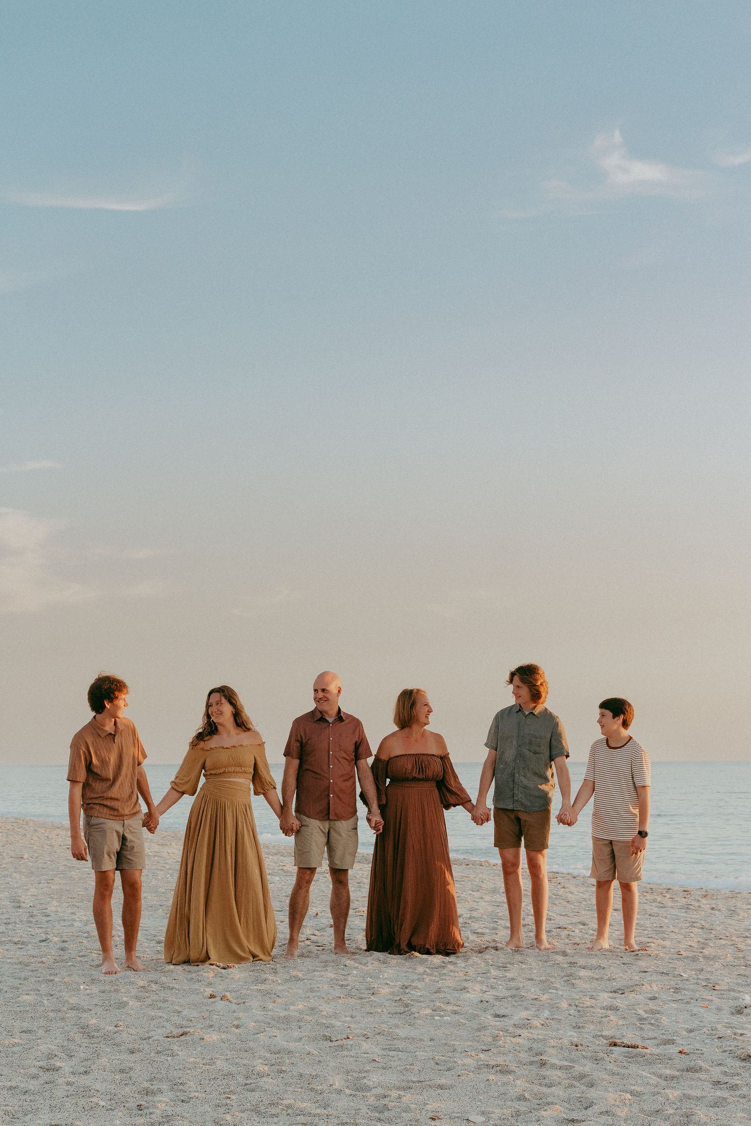 Group of people holding hands while standing together on a white sandy beach at sunset in coordinated earth-tone outfits.