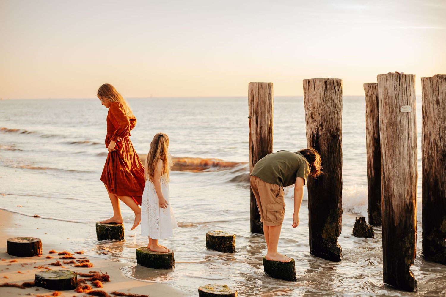 Three people balance on wooden posts along a beach at sunset with waves and golden light in the background.