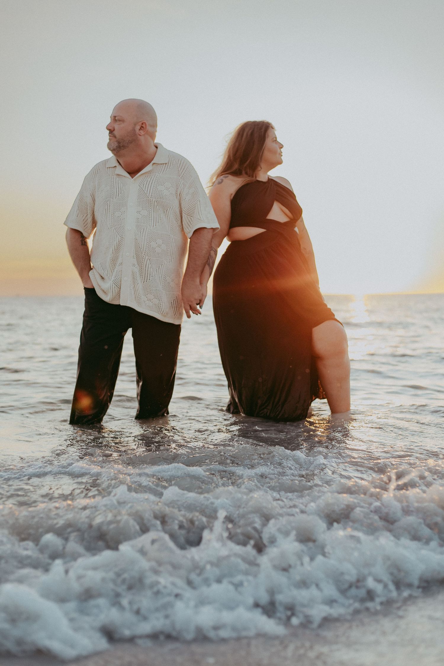 Couple sits together in shallow ocean water at sunset while gentle waves roll in around them.