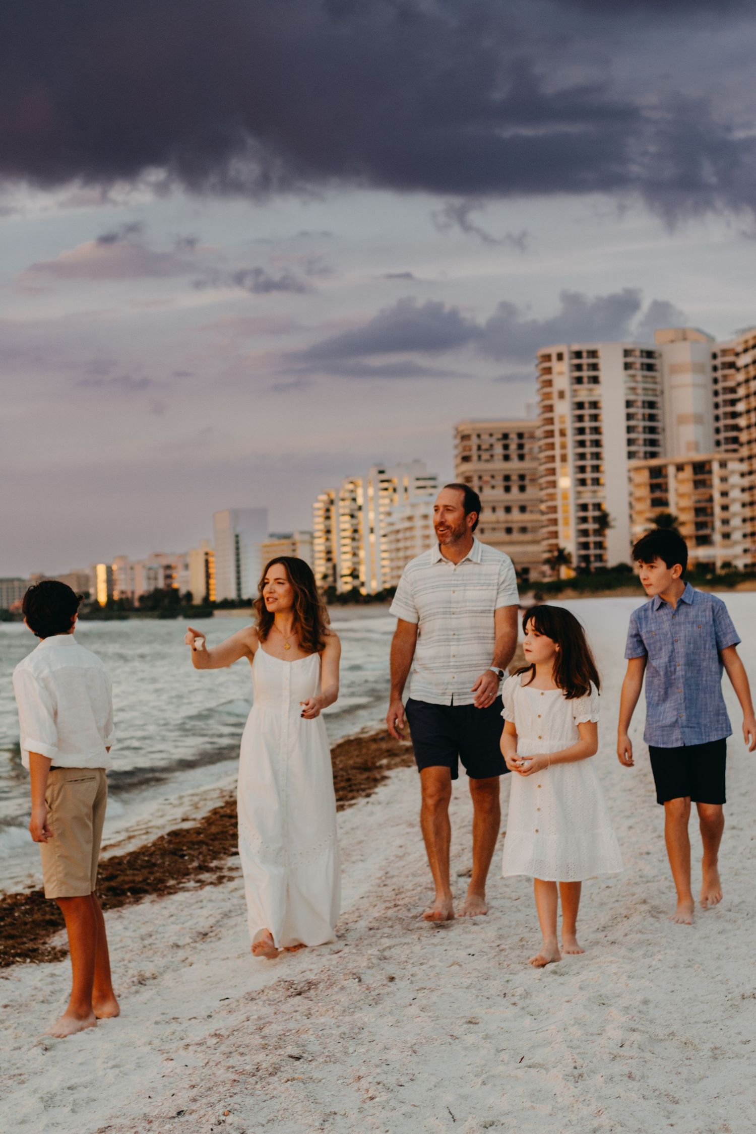 Family enjoying sunset together while walking along a white sandy beach with coastal buildings in background.
